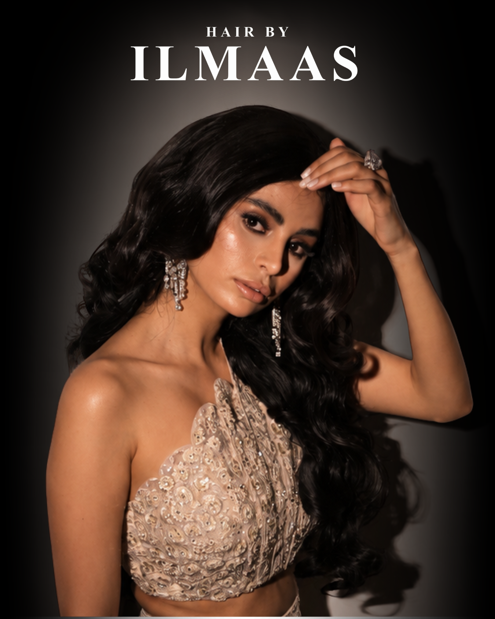 A woman with long wavy dark hair, dressed in a gold sequined top, wearing earrings and a ring, poses against a dark background.