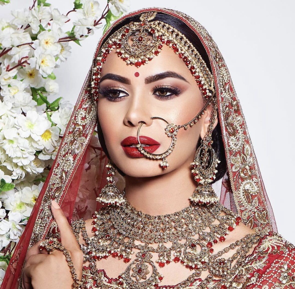 A woman dressed in traditional Indian bridal attire with elaborate jewelry, red and gold embroidery, and a sheer red veil, standing beside white flowers.