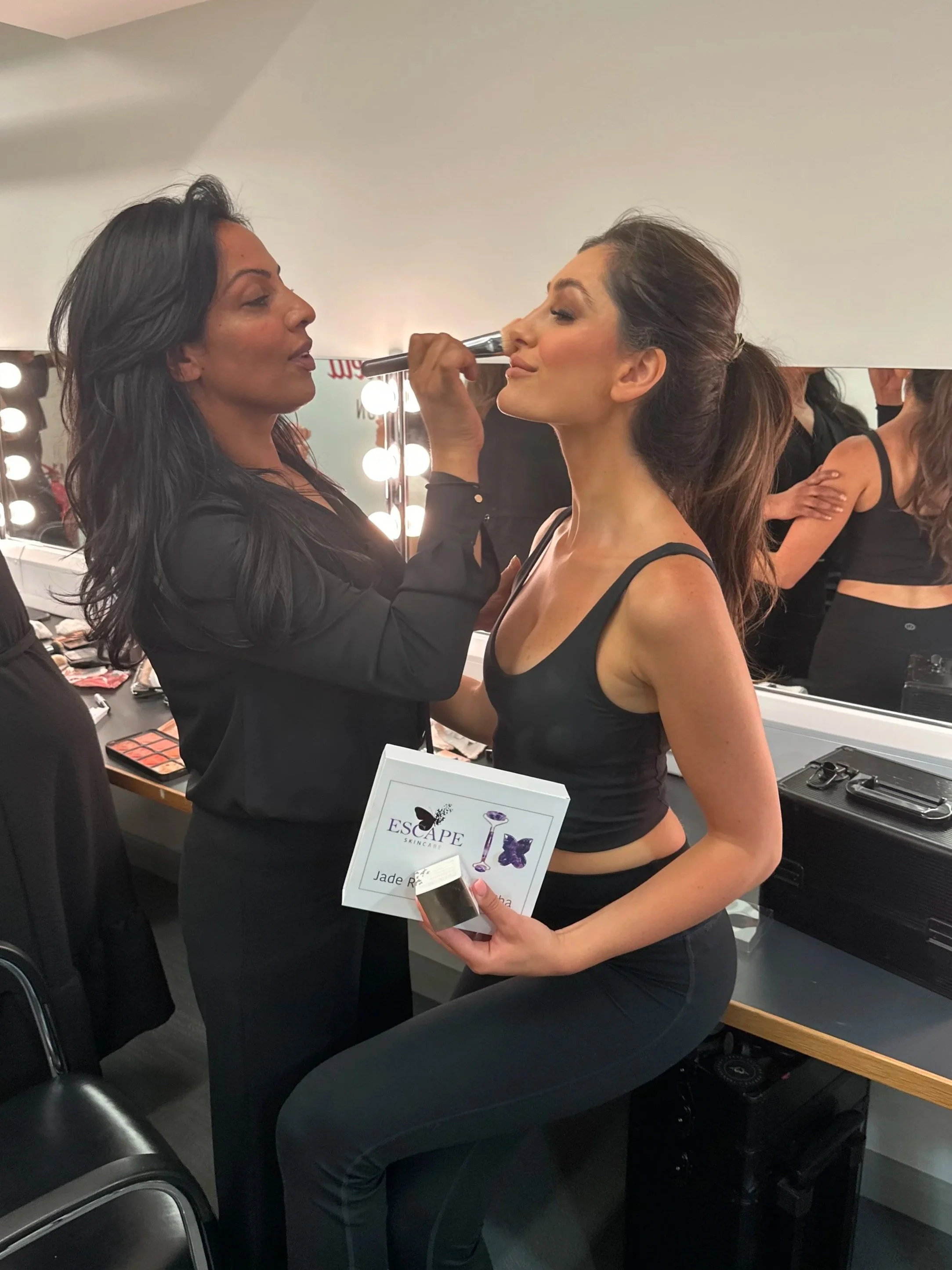 A makeup artist applying makeup to a woman in a black tank top, with a makeup counter and mirror with lights in the background.