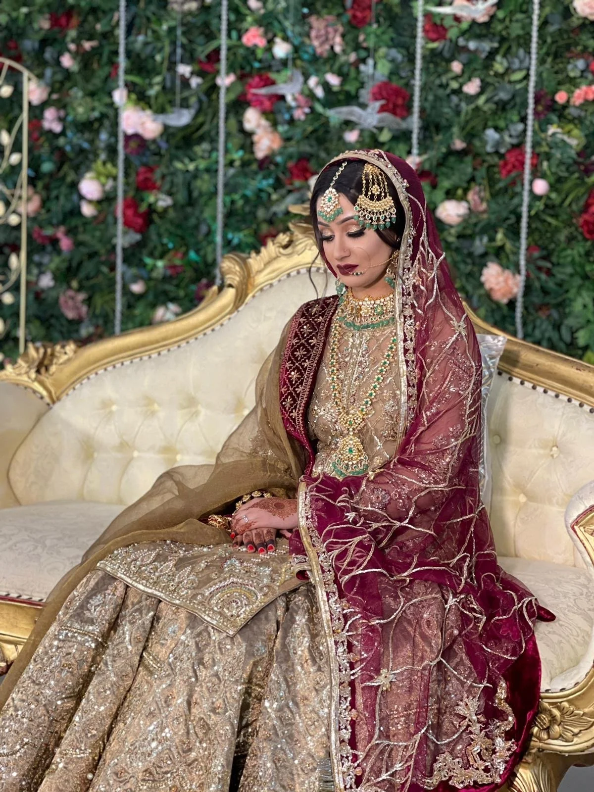 A woman dressed in traditional South Asian bridal attire sitting on a cream and gold ornate sofa with a floral backdrop.