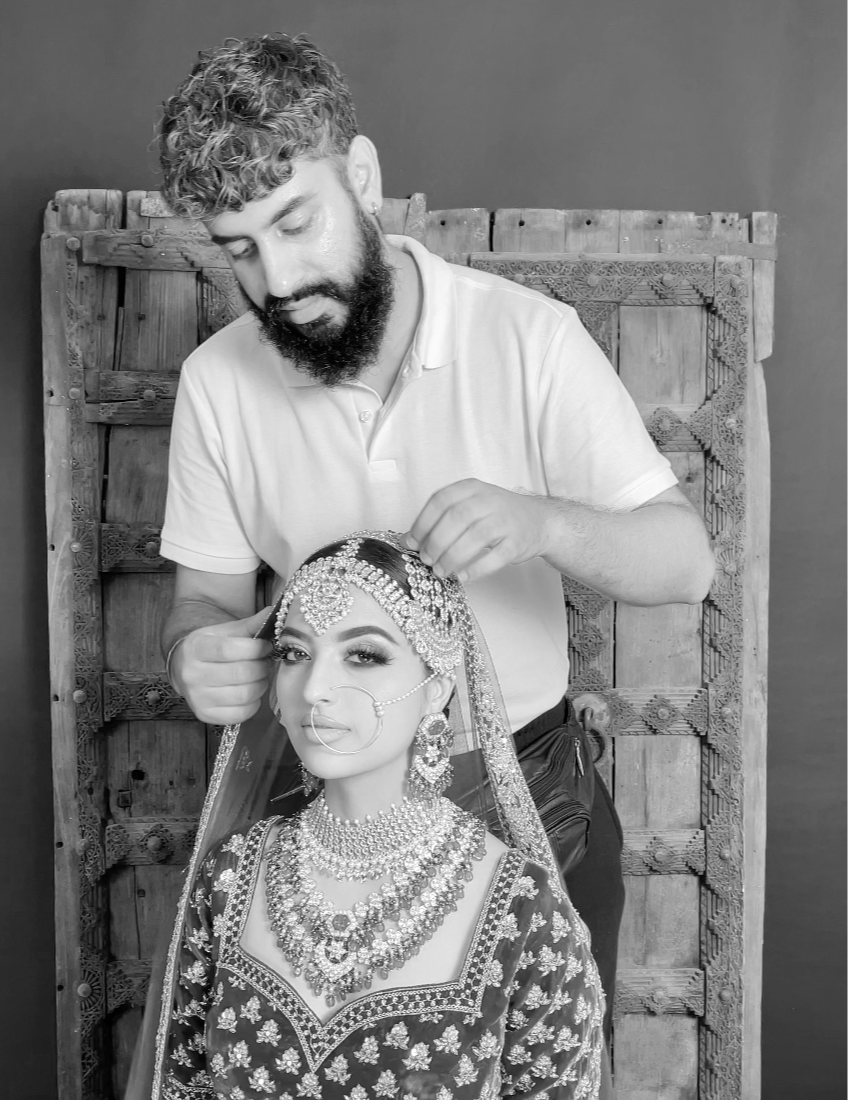 A man in a white polo shirt adjusting the jewelry of a woman in traditional Indian bridal attire, including a heavily embroidered dress, jewelry, nose ring, and veil, against a wooden backdrop.