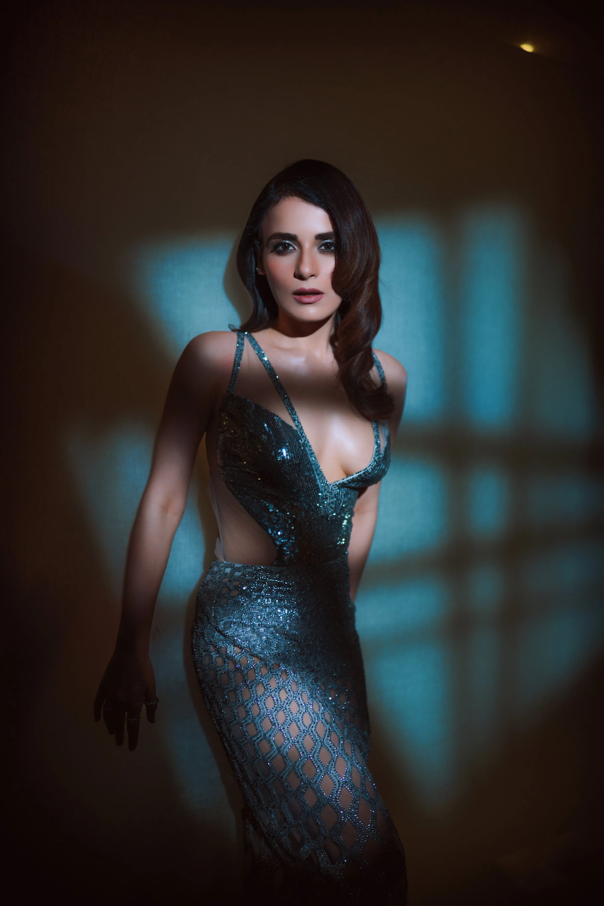 A woman in a sparkly, dark-colored dress with cutouts stands against a softly lit background, looking directly at the camera.