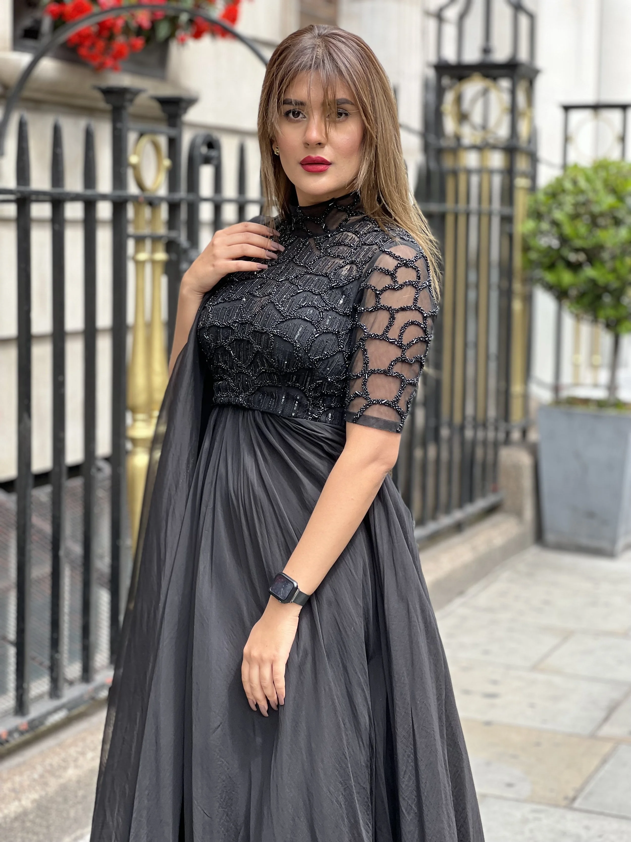 A woman wearing a black dress with intricate lace detailing on the top and semi-sheer sleeves, standing outdoors in front of a metal fence and potted plants.