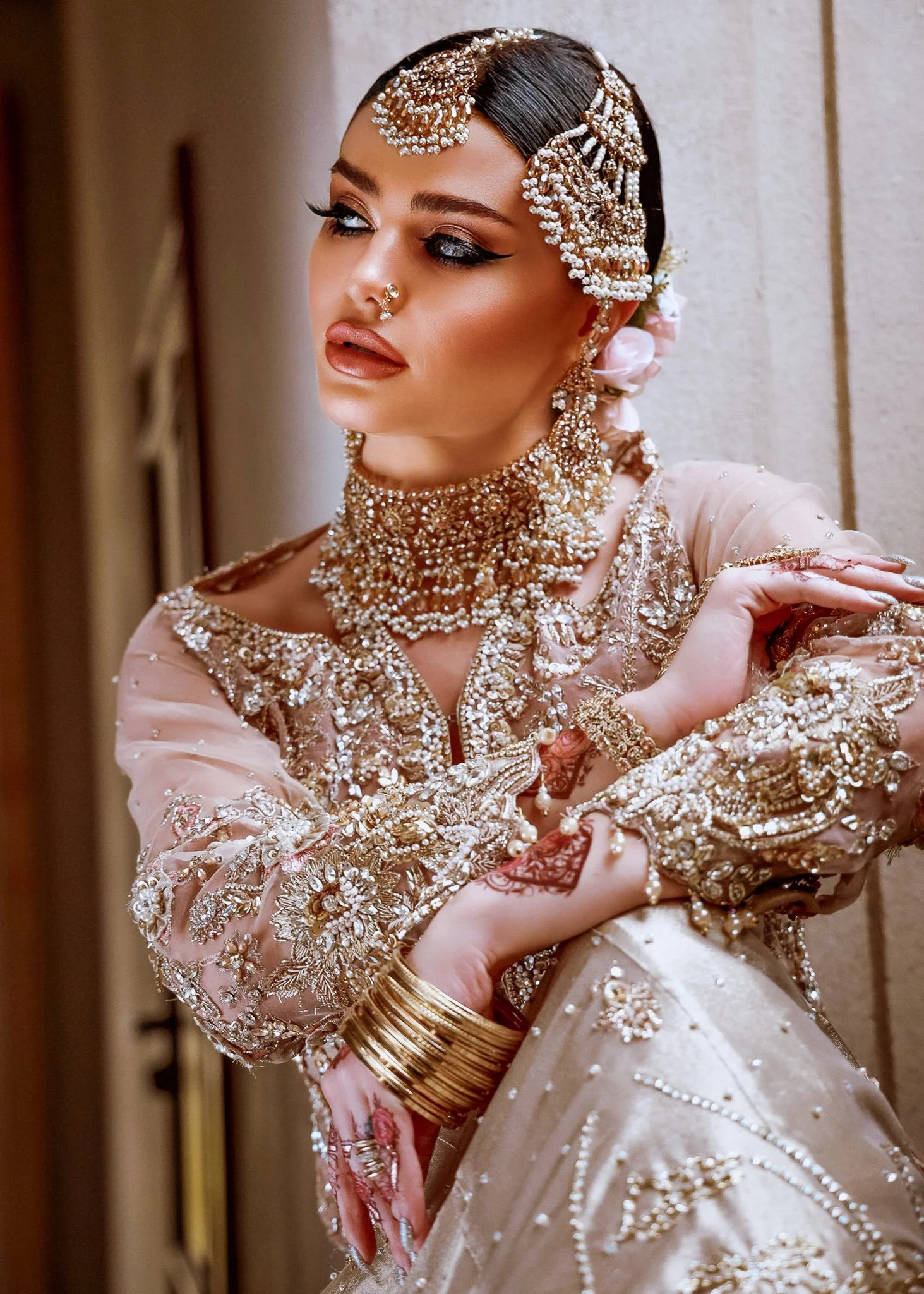 A woman wearing an ornate, heavily embroidered pink and gold traditional wedding dress with elaborate jewelry, including a layered necklace, earrings, and bangles, and a decorative headpiece. She has henna designs on her hands and is posing with her arms crossed.