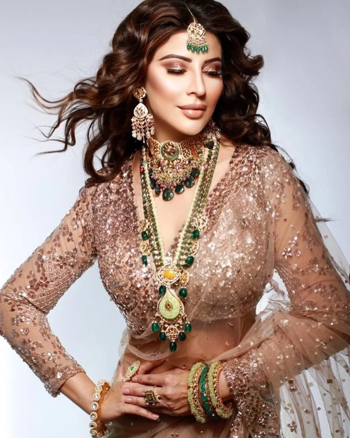A woman with long, curly brown hair wearing traditional Indian jewelry and an embellished, rose gold-colored outfit.