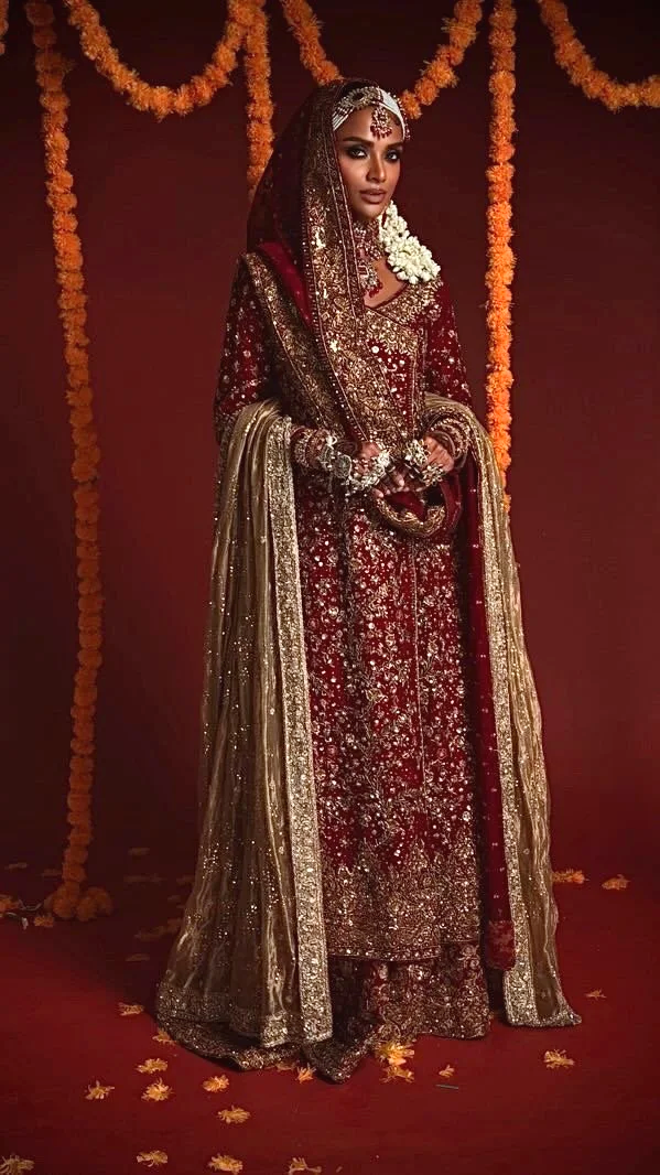 A woman dressed in a traditional red and gold bridal outfit, adorned with extensive jewelry, standing against a decorated backdrop with orange marigold flower garlands.