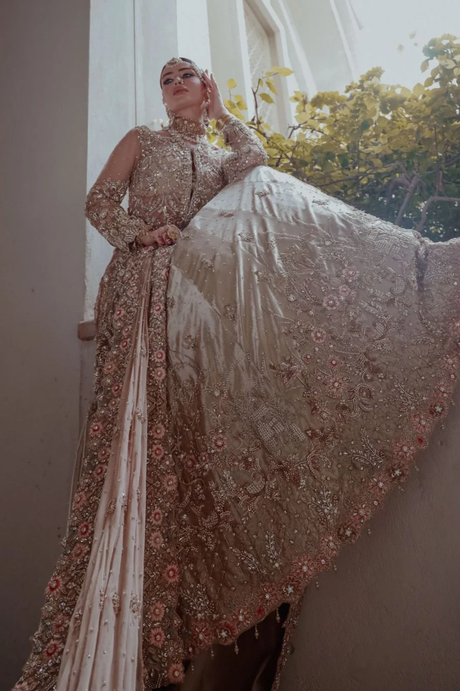A woman dressed in an ornate, embroidered gown with detailed beadwork and embellishments, posing indoors near a window with sunlight and greenery outside.