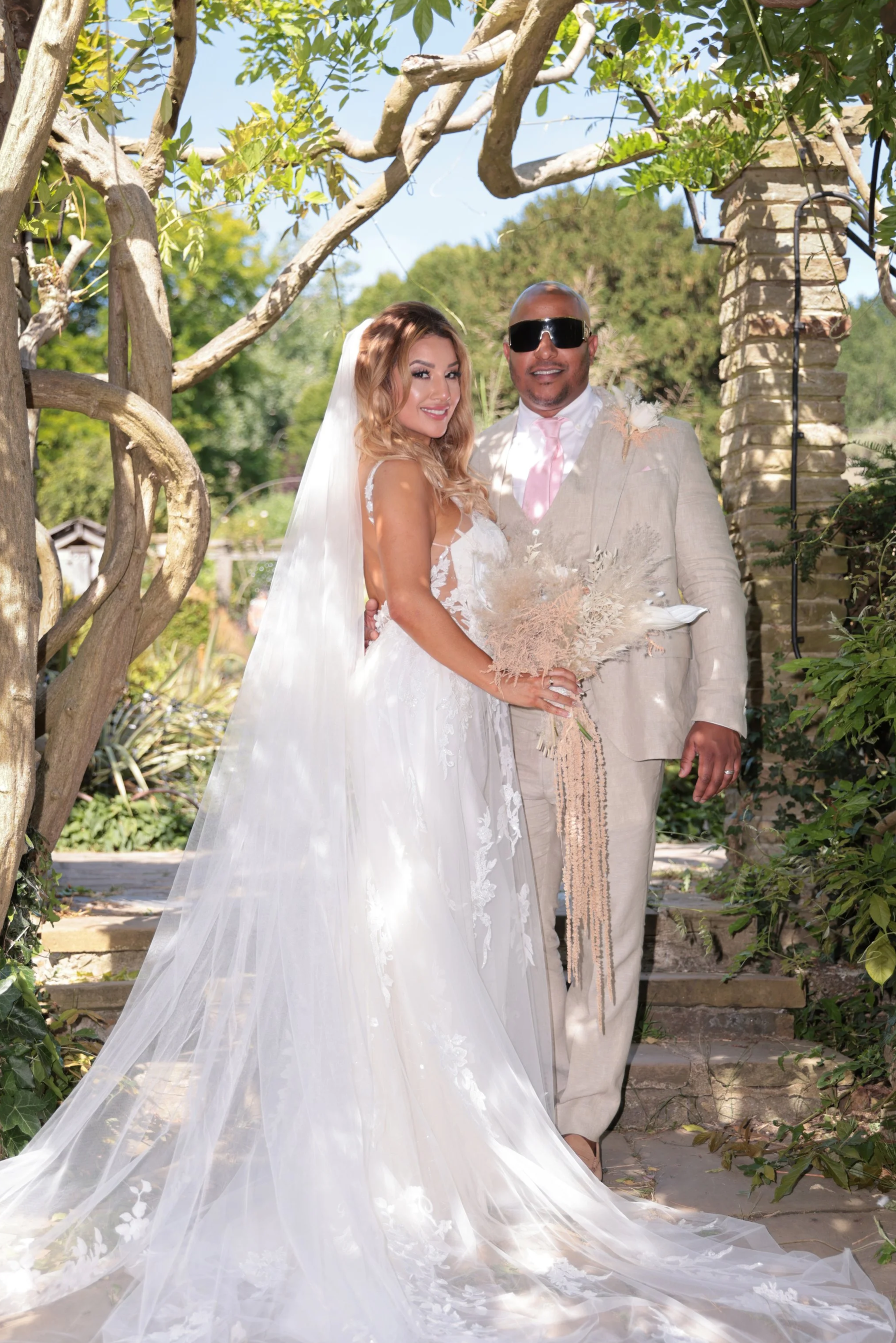 Bride in a white wedding gown and veil standing next to groom in a beige suit with sunglasses, holding a bouquet of dried flowers outdoors.