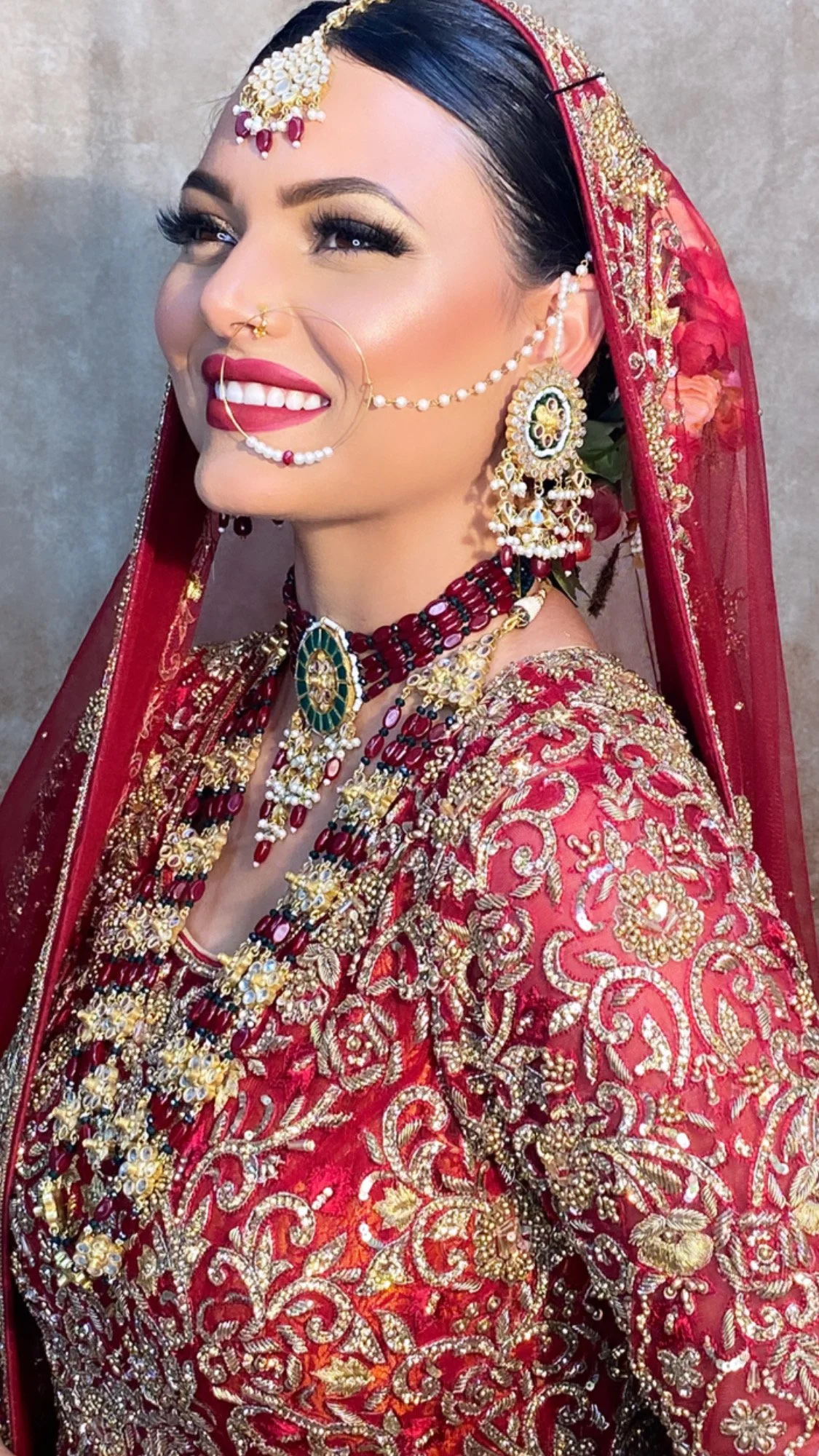 A woman dressed in traditional Indian bridal attire, wearing a red heavily embroidered wedding saree with jewelry, including earrings, necklaces, and a headpiece.