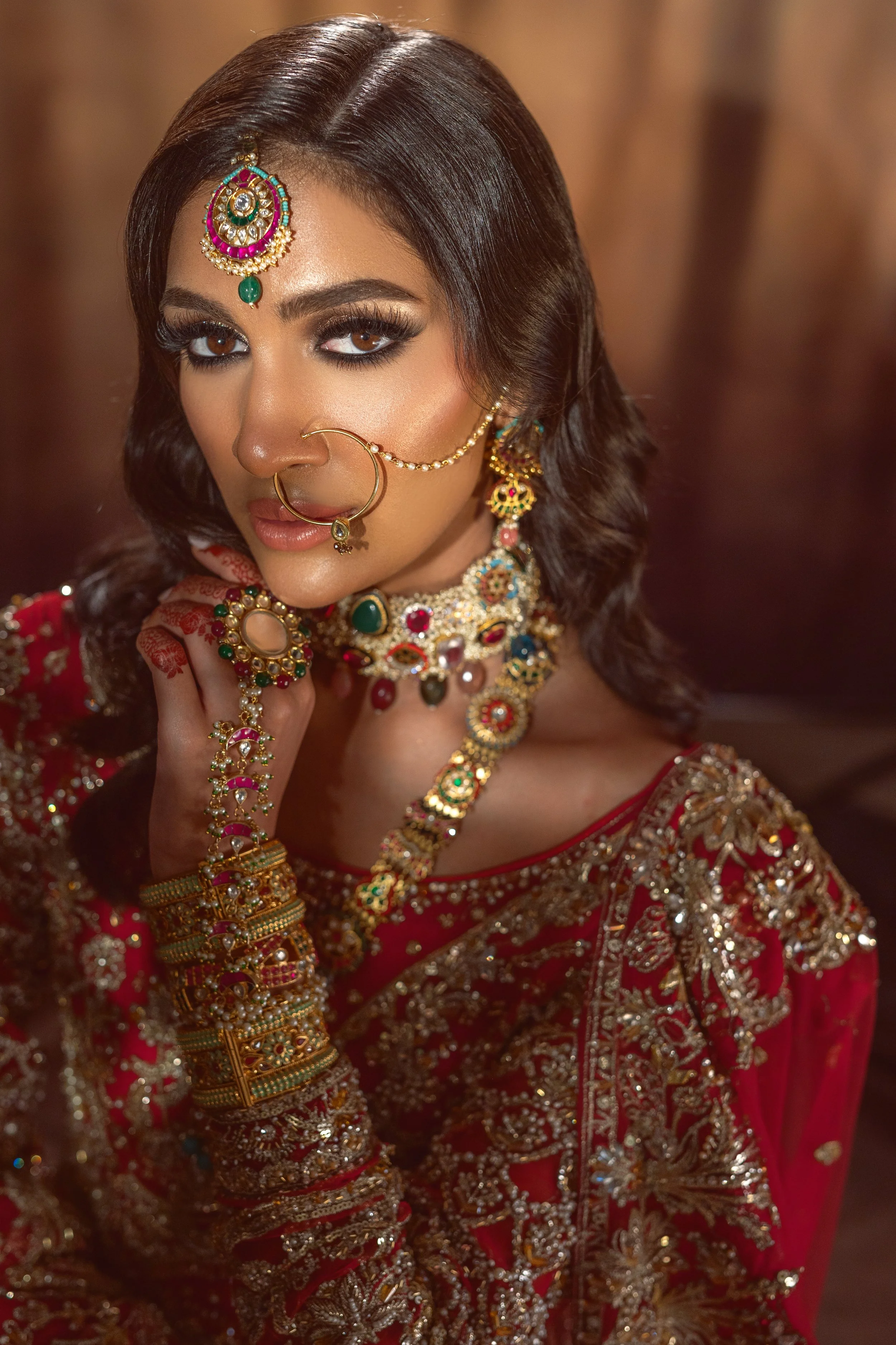 A woman wearing traditional Indian bridal attire and jewelry, including a red embroidered saree, heavy necklaces, earrings, nose ring, and bangles, with henna on her hand and dark, wavy hair.