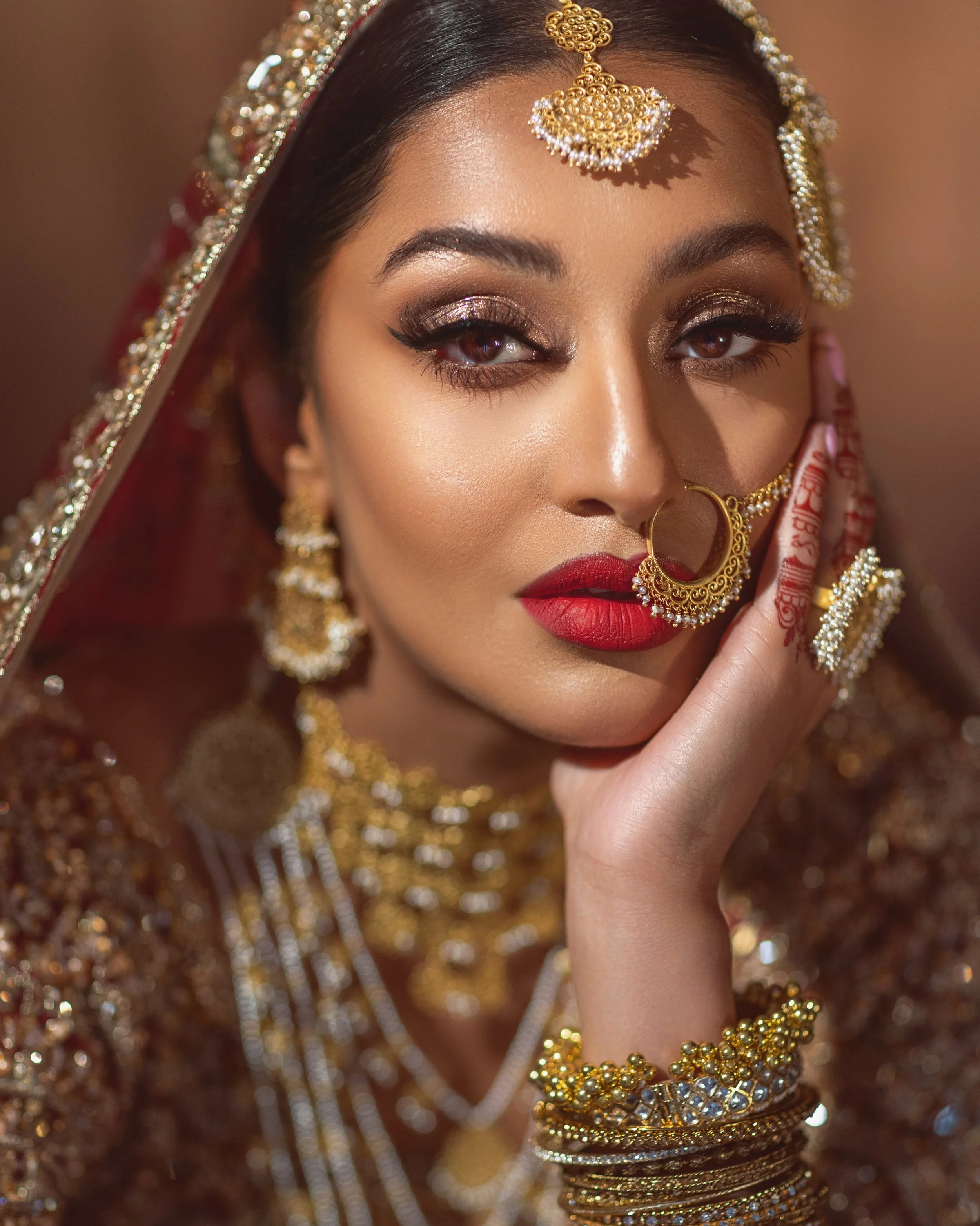 A woman dressed in traditional Indian jewelry and attire, with elaborate gold jewelry, including earrings, nose ring, necklace, and bangles, and wearing bold makeup with red lipstick and shimmery eye makeup.