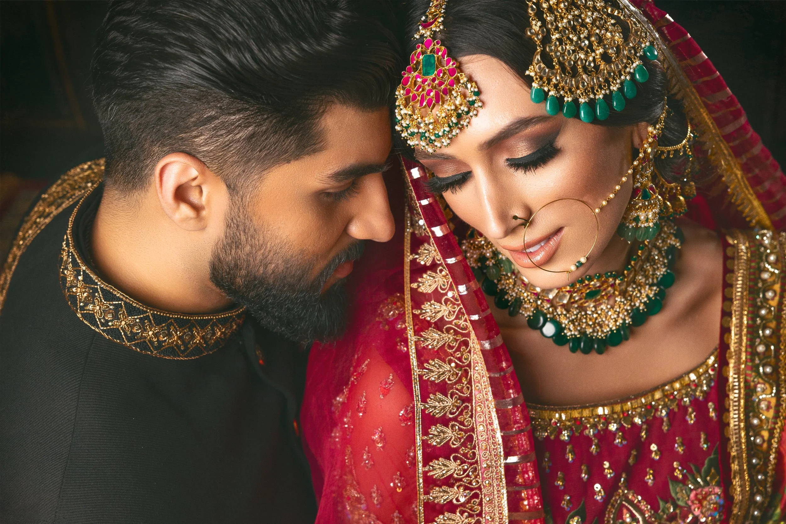 A bride and groom with their foreheads touching, dressed in traditional Indian wedding attire. The bride wears an ornate red and gold saree, heavily adorned with jewelry including earrings, a nose ring, necklaces, and a headpiece. The groom wears a black outfit with gold embroidery on the collar.