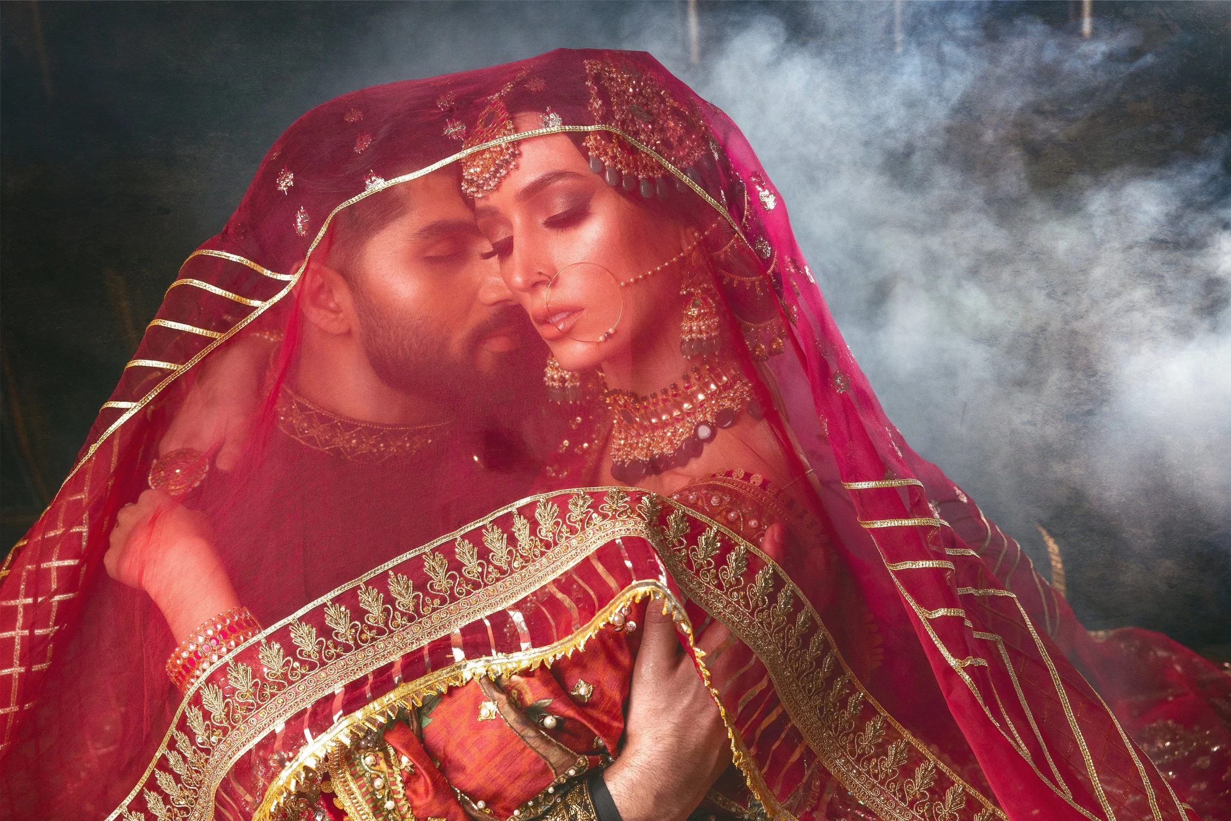 A couple dressed in traditional Indian wedding attire, with the bride in red and gold jewelry and veil, and the groom in matching attire, embracing with eyes closed amid a smoky atmosphere.