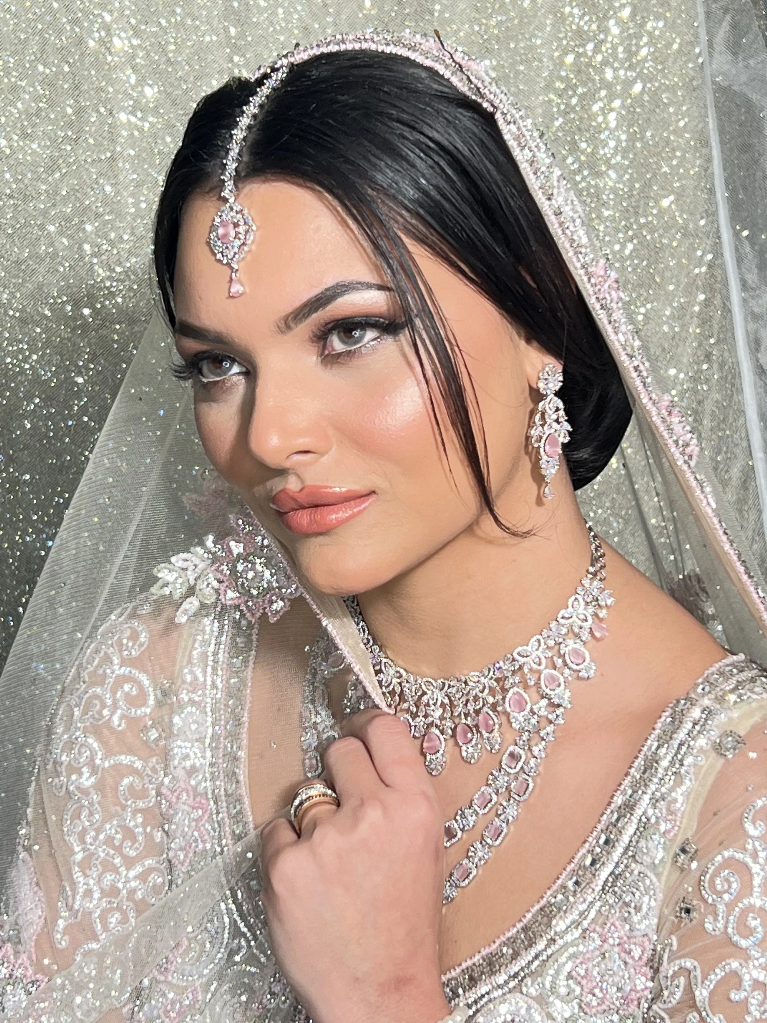 A woman wearing a bridal veil and intricate jewelry, including earrings, a necklace, and a headpiece, with makeup and dark hair styled elegantly for a wedding.