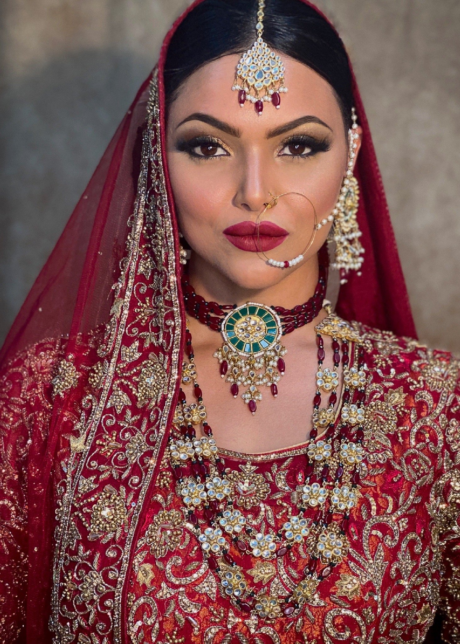 A woman in traditional Indian bridal attire with intricate gold and red embroidery, wearing ornate jewelry including a maang tikka, nose ring, multiple necklaces, earrings, and bangles.