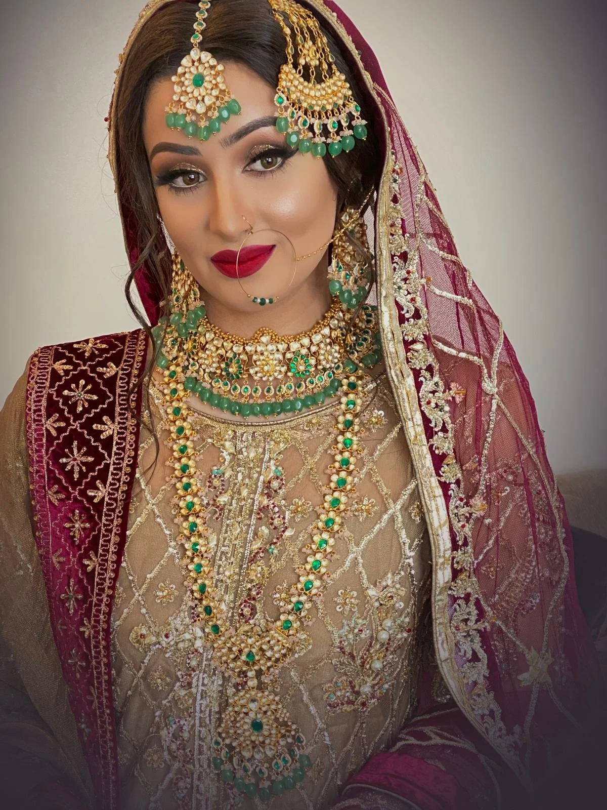 A woman dressed in traditional Indian bridal attire with intricate jewelry, including a maang tikka, earrings, necklaces, nose ring, and bangles, wearing a richly embroidered maroon and gold outfit with a matching veil.