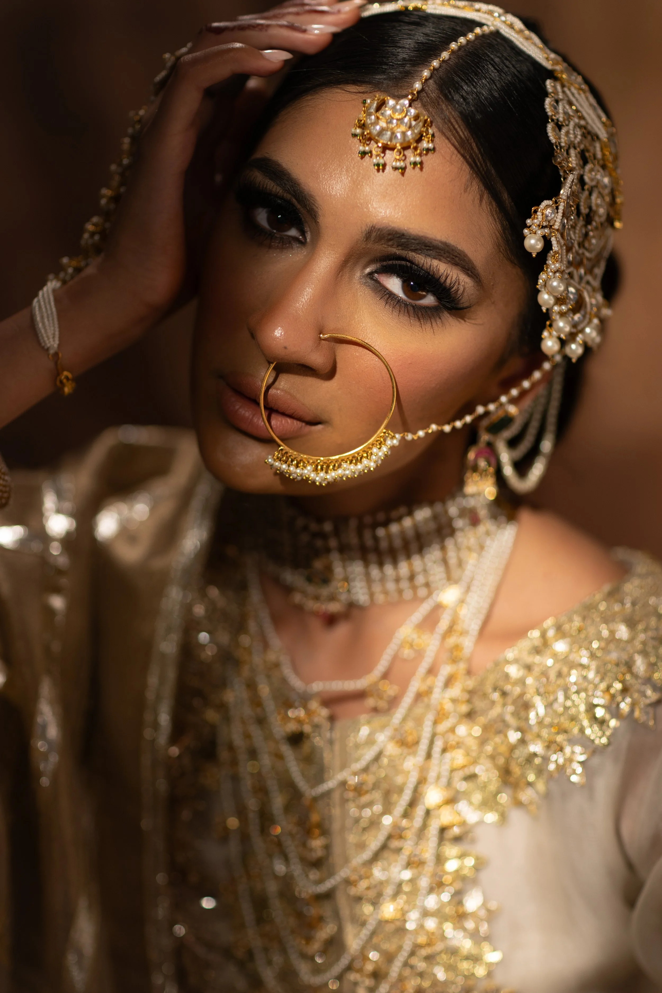 A woman dressed in traditional Indian bridal attire with intricate gold jewelry, including a nose ring, earrings, necklace, and headpiece. She has makeup with bold eye makeup and is posing with her hand on her head.