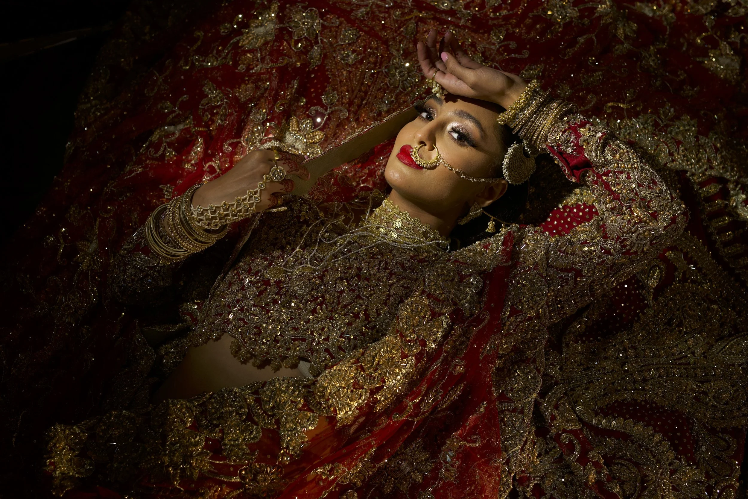 A woman dressed in traditional bridal attire with ornate gold jewelry and embroidery, lying on a richly decorated red and gold fabric.
