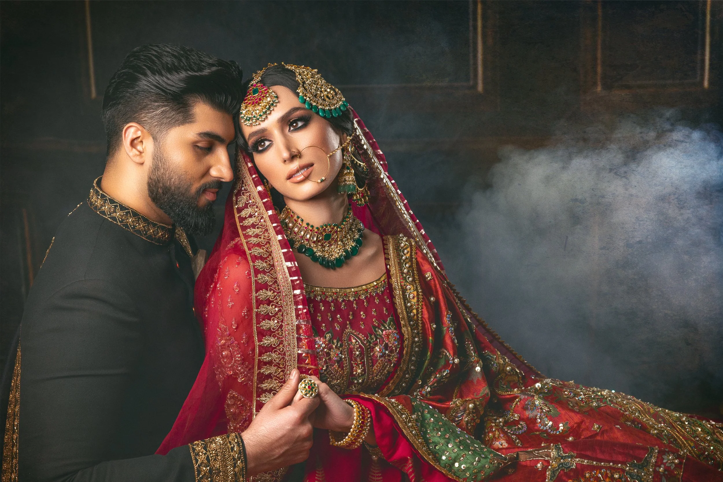 A couple dressed in traditional Indian wedding attire, the man in black with gold embroidery and the woman in a richly adorned red and green saree with elaborate jewelry, including a necklace, earrings, and headpiece.