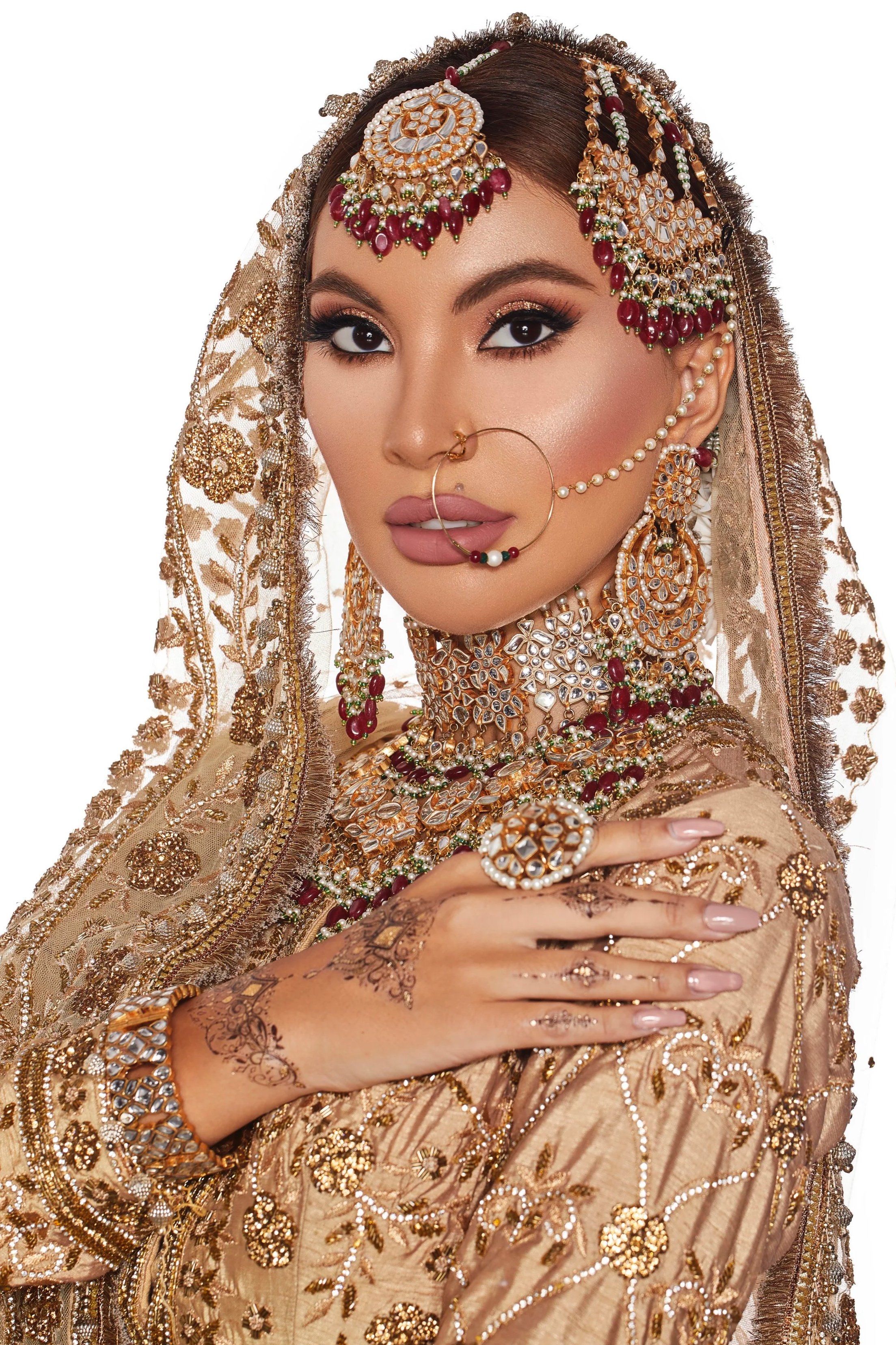 A woman dressed in traditional Indian bridal attire, wearing gold and jeweled jewelry, including a headpiece, earrings, a nose ring, multiple necklaces, and rings. She has henna designs on her hand and is posing with one hand on her shoulder.