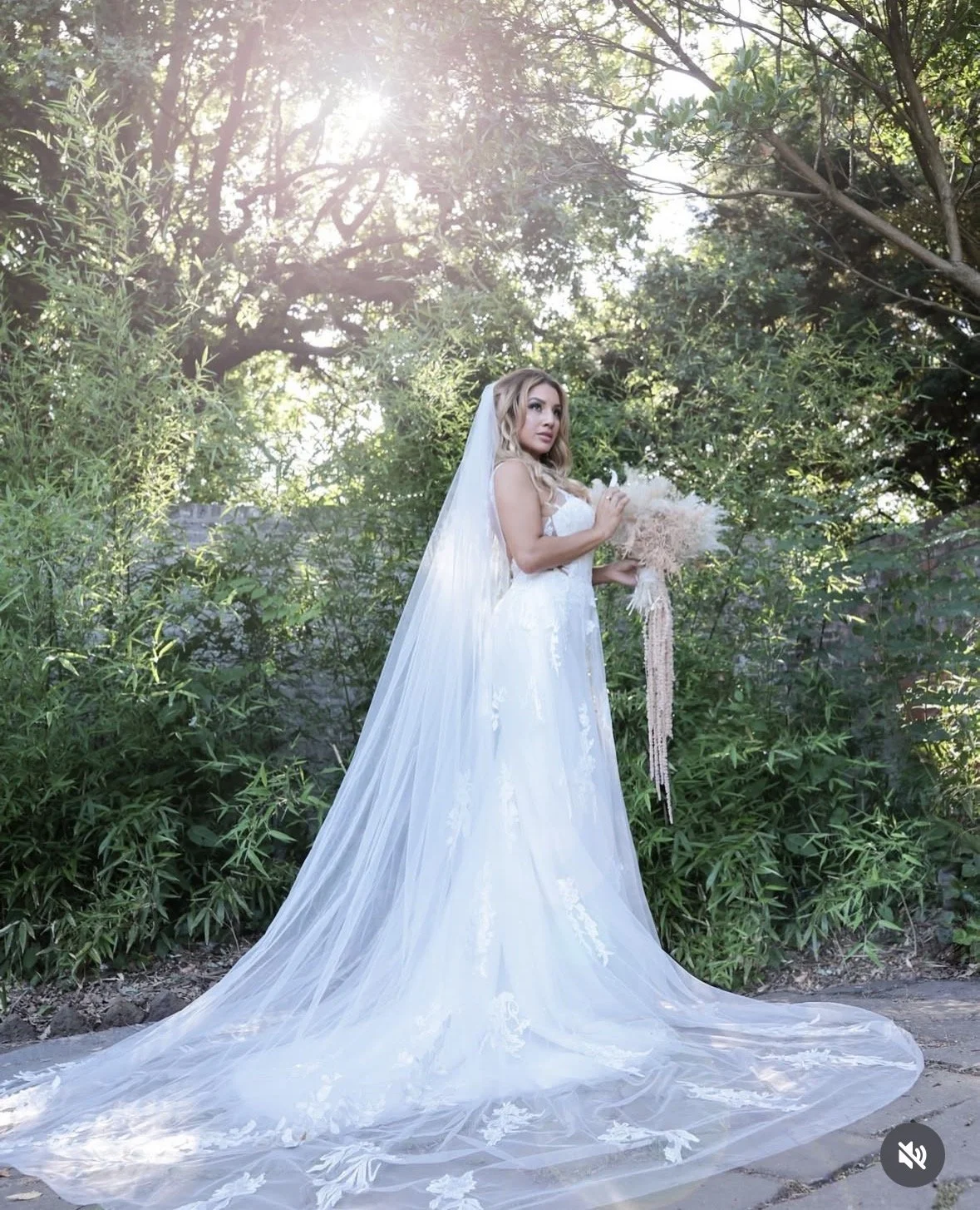 A bride in a white wedding dress holding a bouquet outdoors with lush greenery and trees, sunlight shining through the leaves.