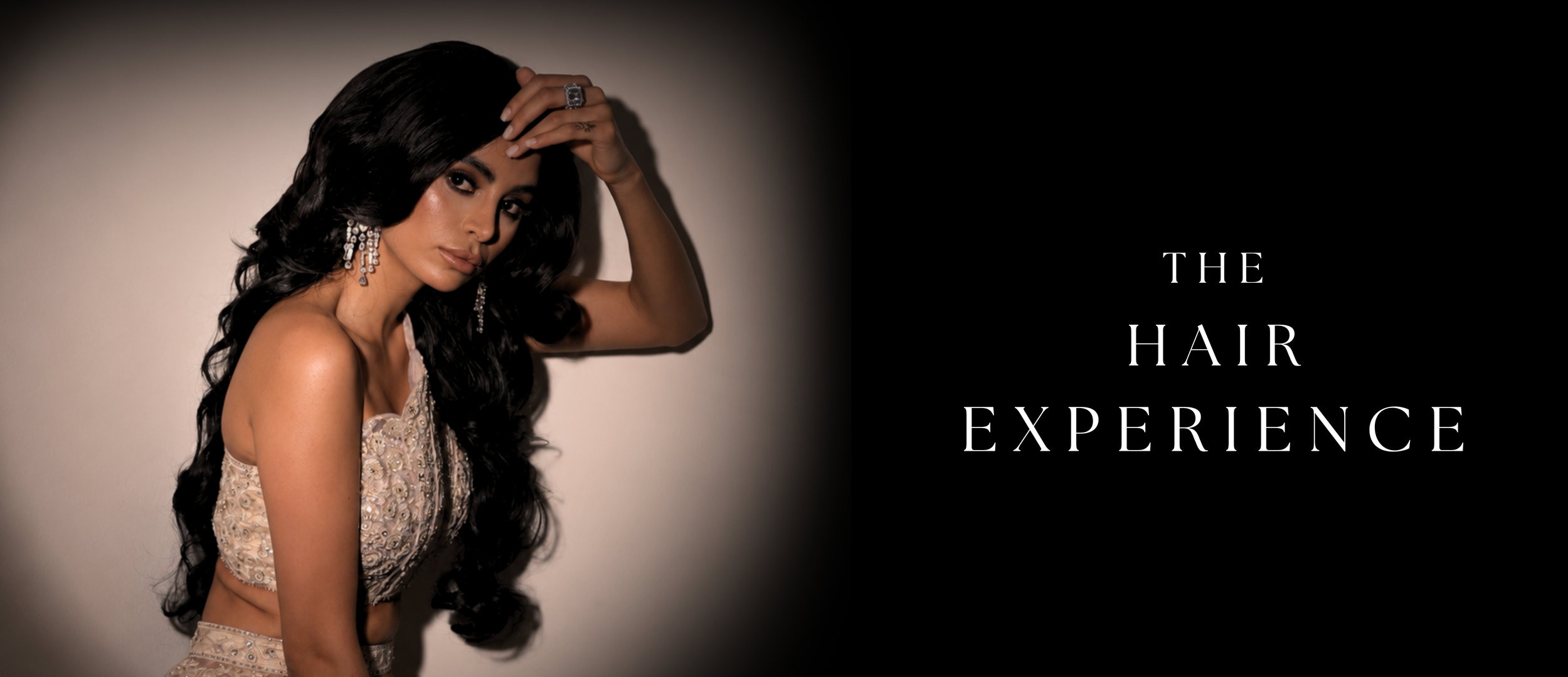 A woman with long black hair and jewelry, wearing an elegant dress, touching her head, beside the words 'The Hair Experience' on a black background.
