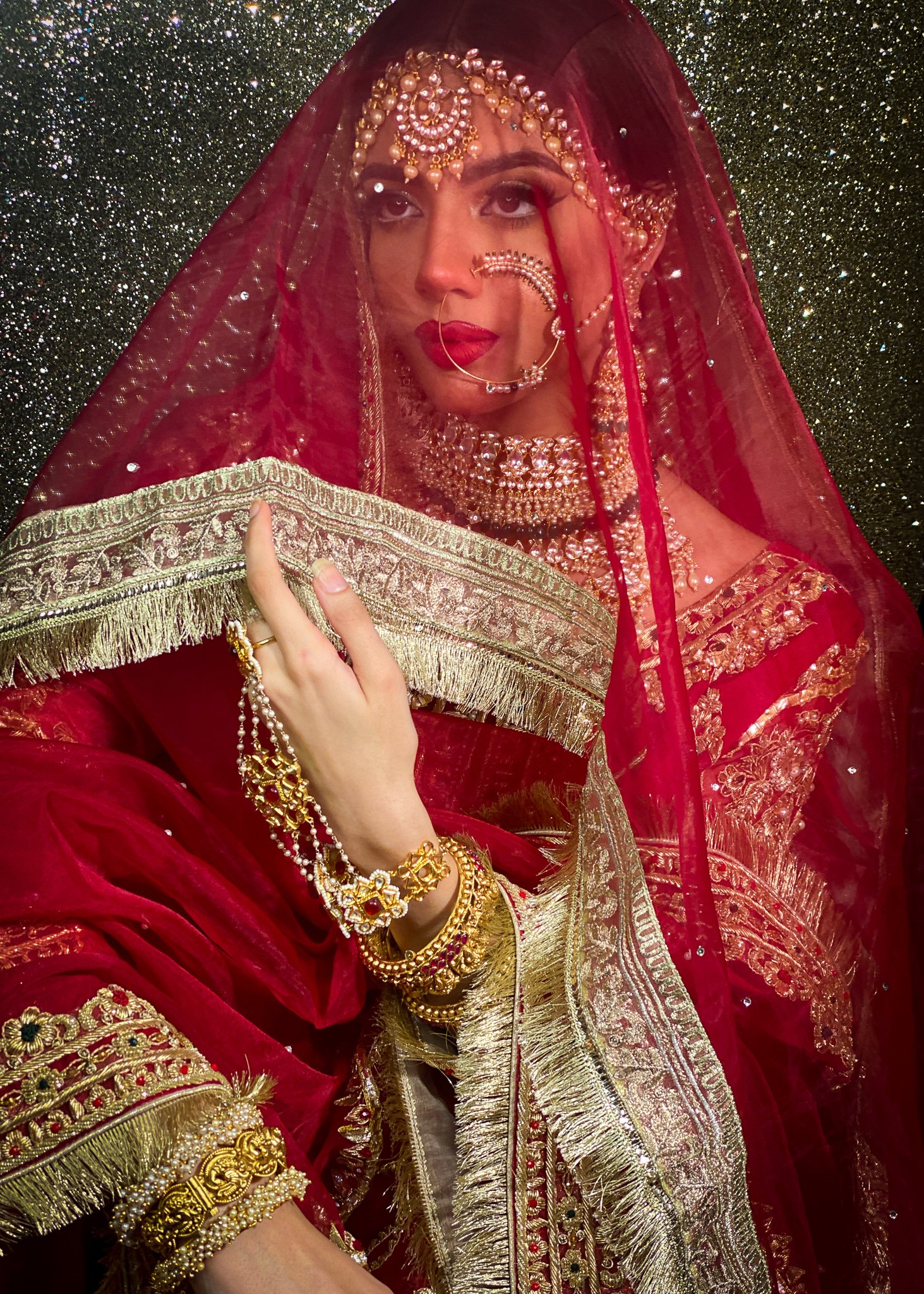 A woman dressed in traditional Indian bridal attire with gold jewelry, red and gold embroidery, and a red veil, holding a silver and gold embroidered cloth in front of a sparkling black background.