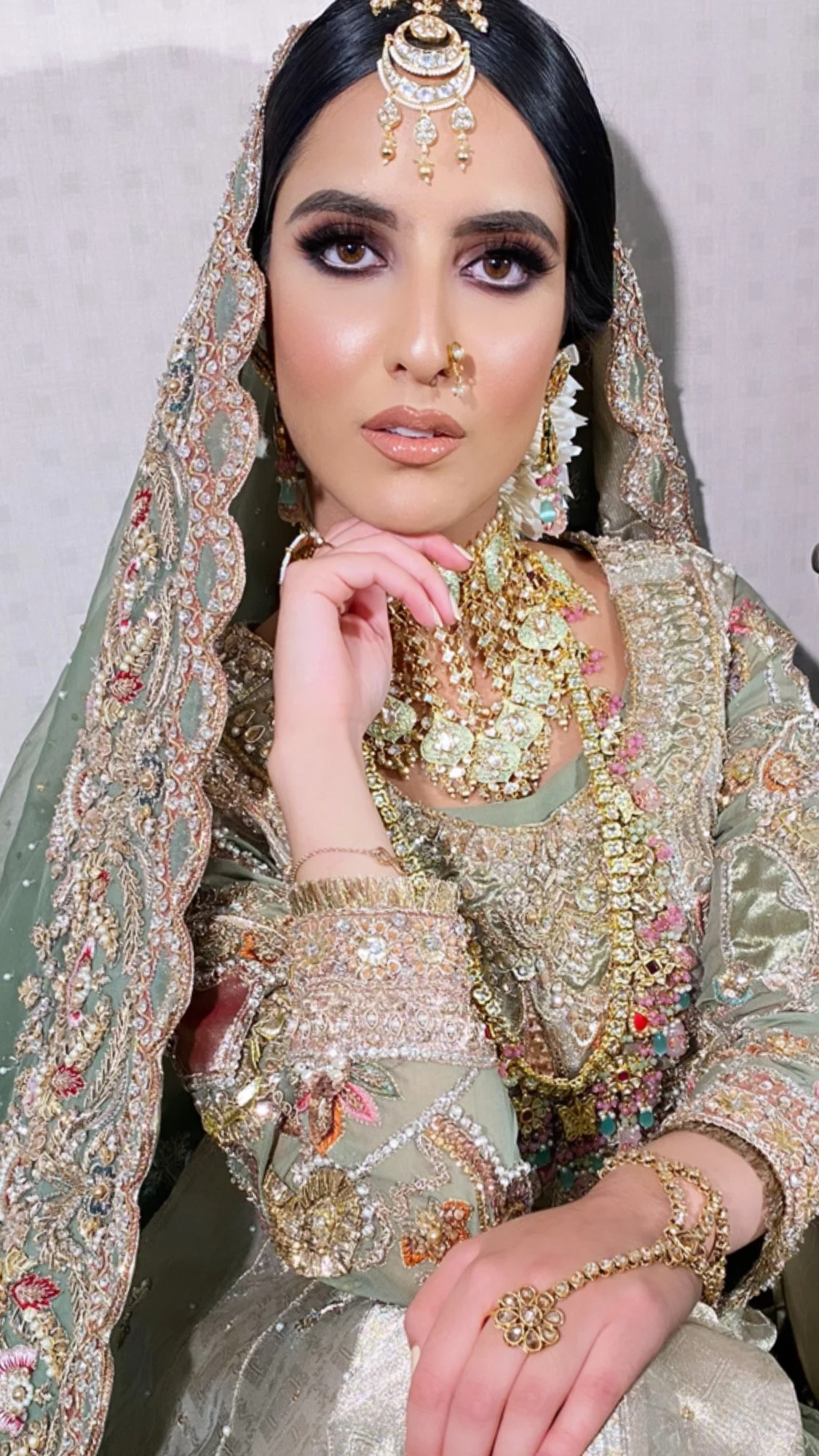 A woman dressed in traditional South Asian bridal attire with intricate embroidery, wearing a heavily embellished greenish gold outfit, adorned with numerous gold and gemstone jewelry including a headpiece, earrings, necklaces, rings, and bracelets.