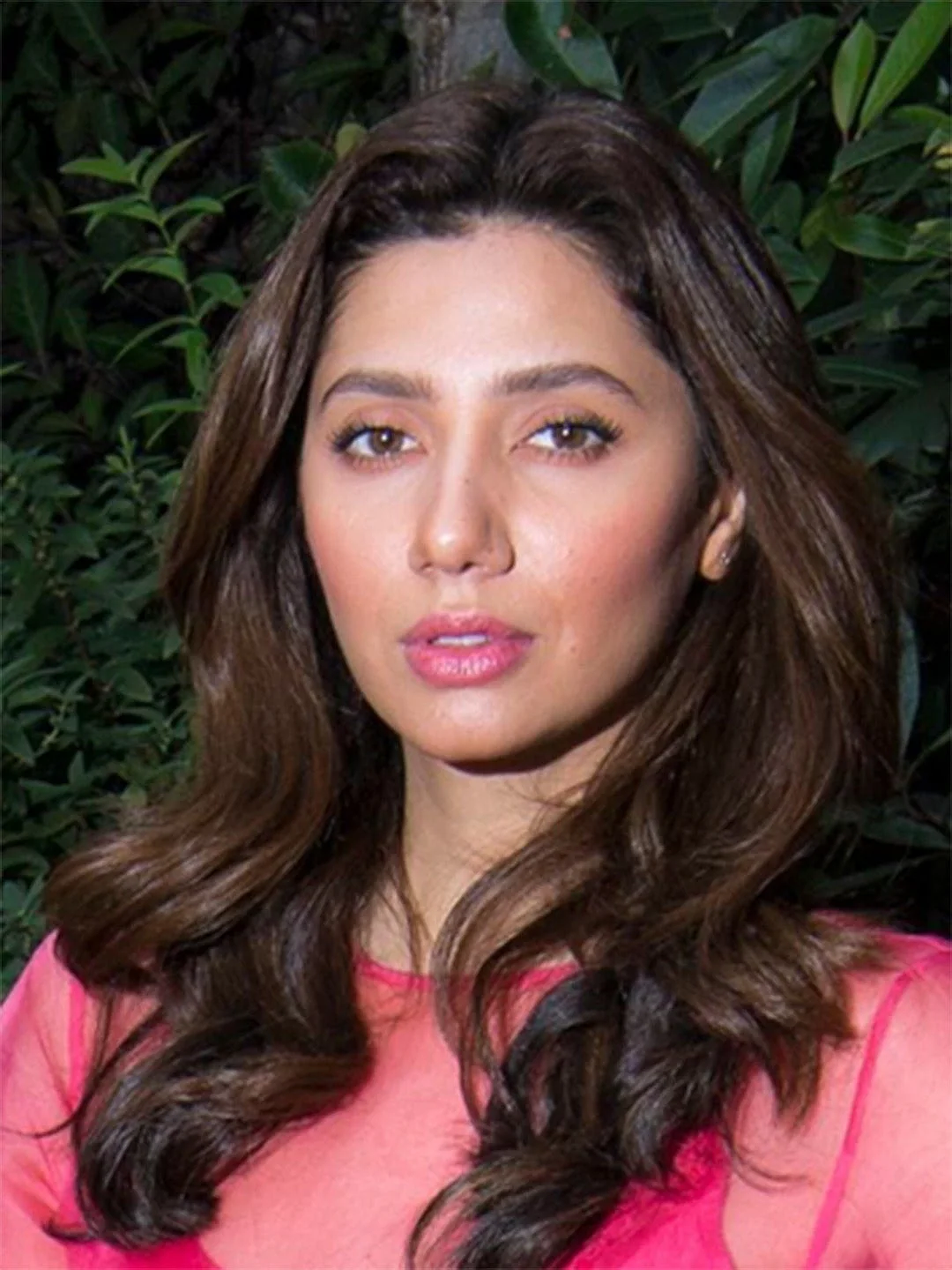 A woman with wavy brown hair and light makeup, wearing a pink top, standing in front of green foliage.
