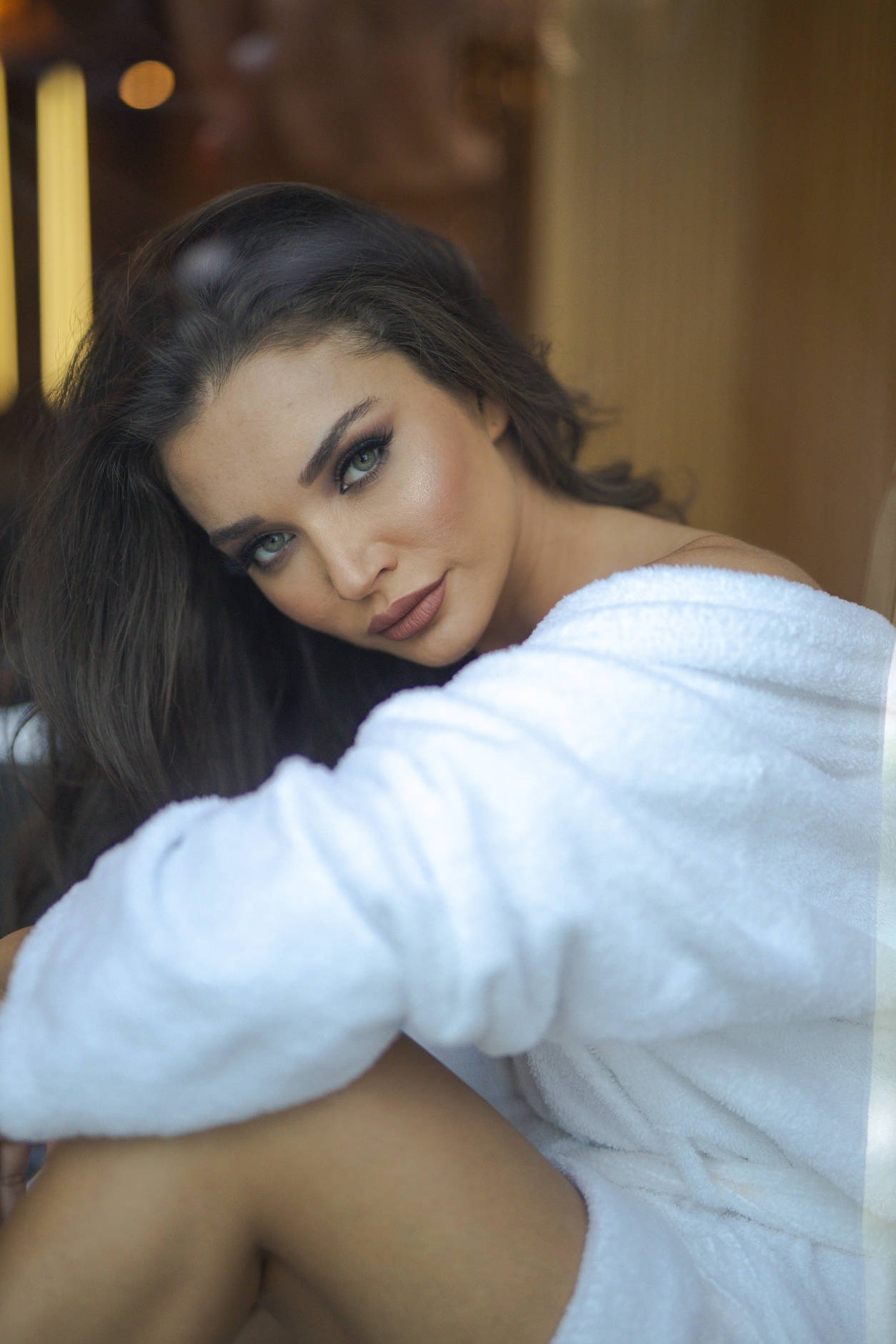 A woman with blue eyes and dark hair, wearing makeup, resting her head on her knee, wearing a white towel.