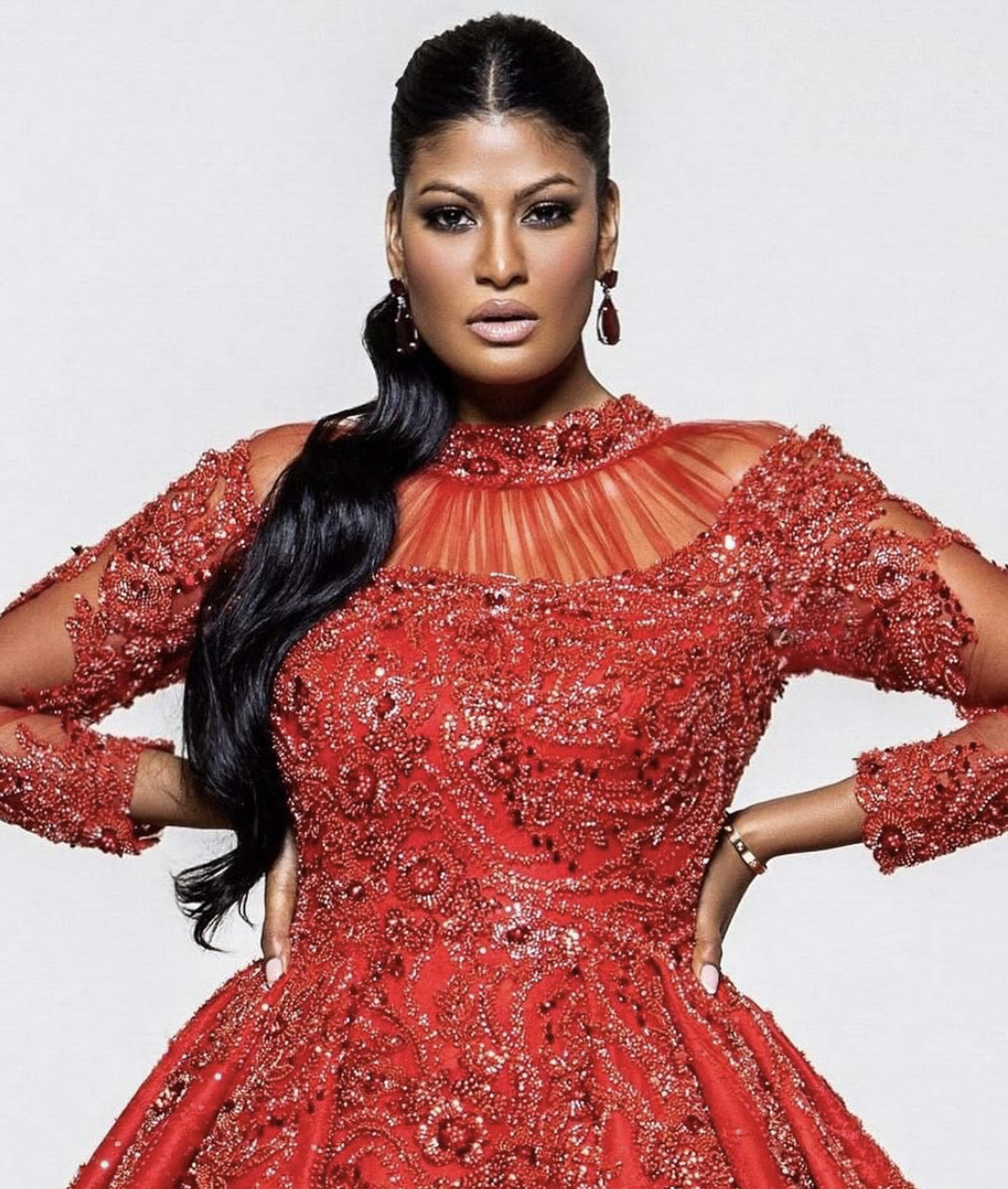 A woman with long black hair in a side ponytail, wearing a red, heavily embellished gown with intricate beadwork and lace details, and matching earrings.