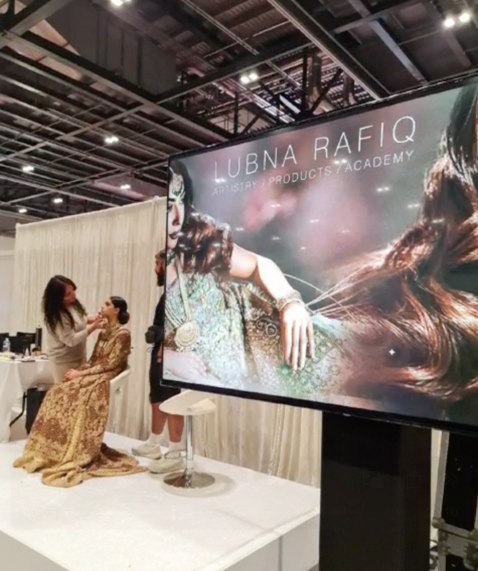 A beauty artist or stylist applying makeup or performing a grooming procedure on a seated woman dressed in a traditional gold and bronze patterned outfit at a cosmetology or grooming event, with a large sign that reads 'LUBNA RAFIQ ARTISTRY PRODUCTS ACADEMY' and a backdrop of a curtain.