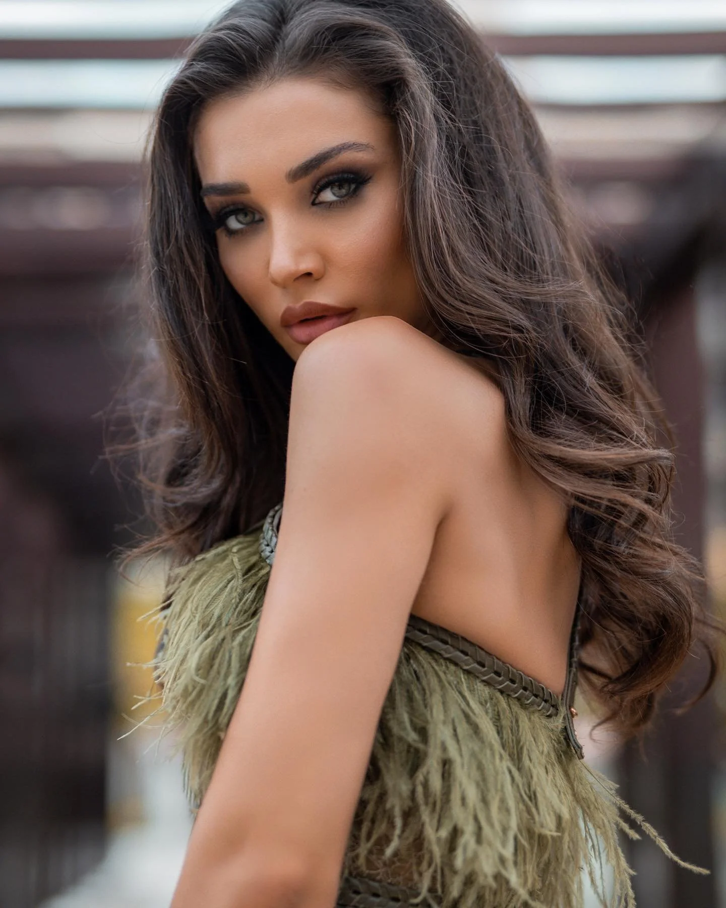 A woman with long, wavy brown hair and makeup, looking over her shoulder outdoors in front of a blurred background.