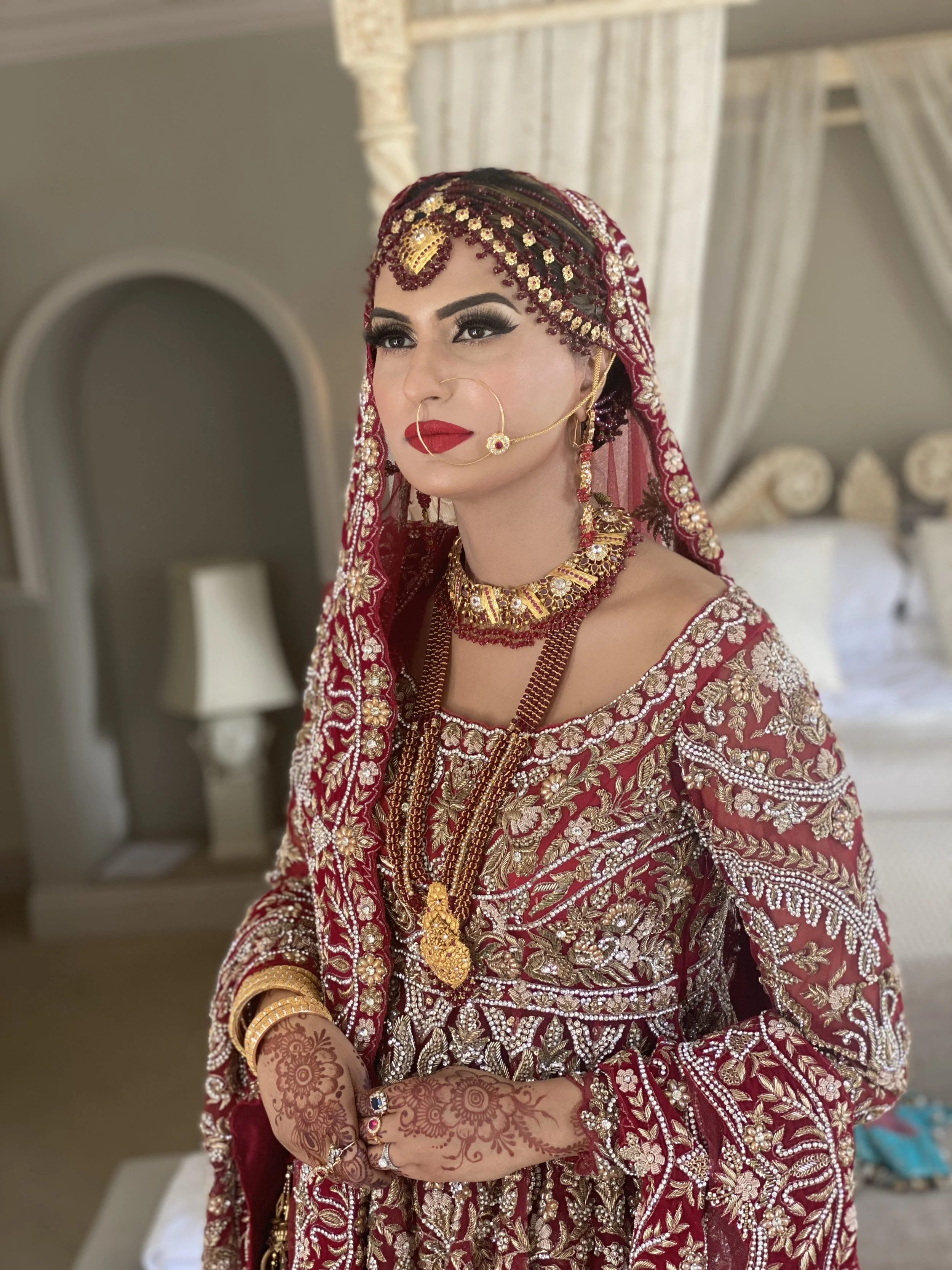 A woman dressed in traditional South Asian bridal attire, adorned with intricate jewelry, henna, and a richly embroidered red and gold outfit, standing in a decorated room.