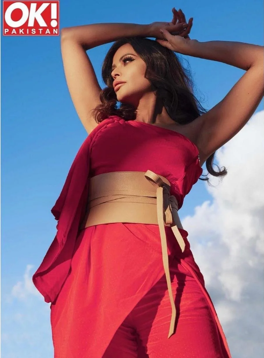 A woman with dark hair wearing a red outfit with a beige belt, posing outdoors against a blue sky with white clouds.