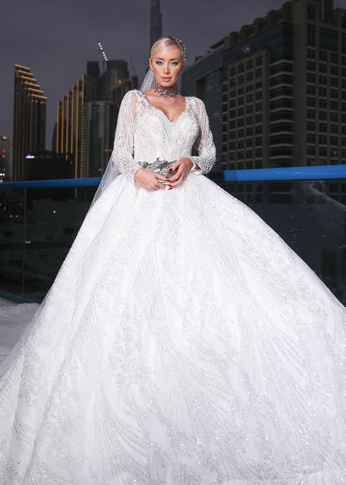 A woman in a white wedding dress with intricate lace and beadwork, standing outdoors against a cityscape background at dusk.