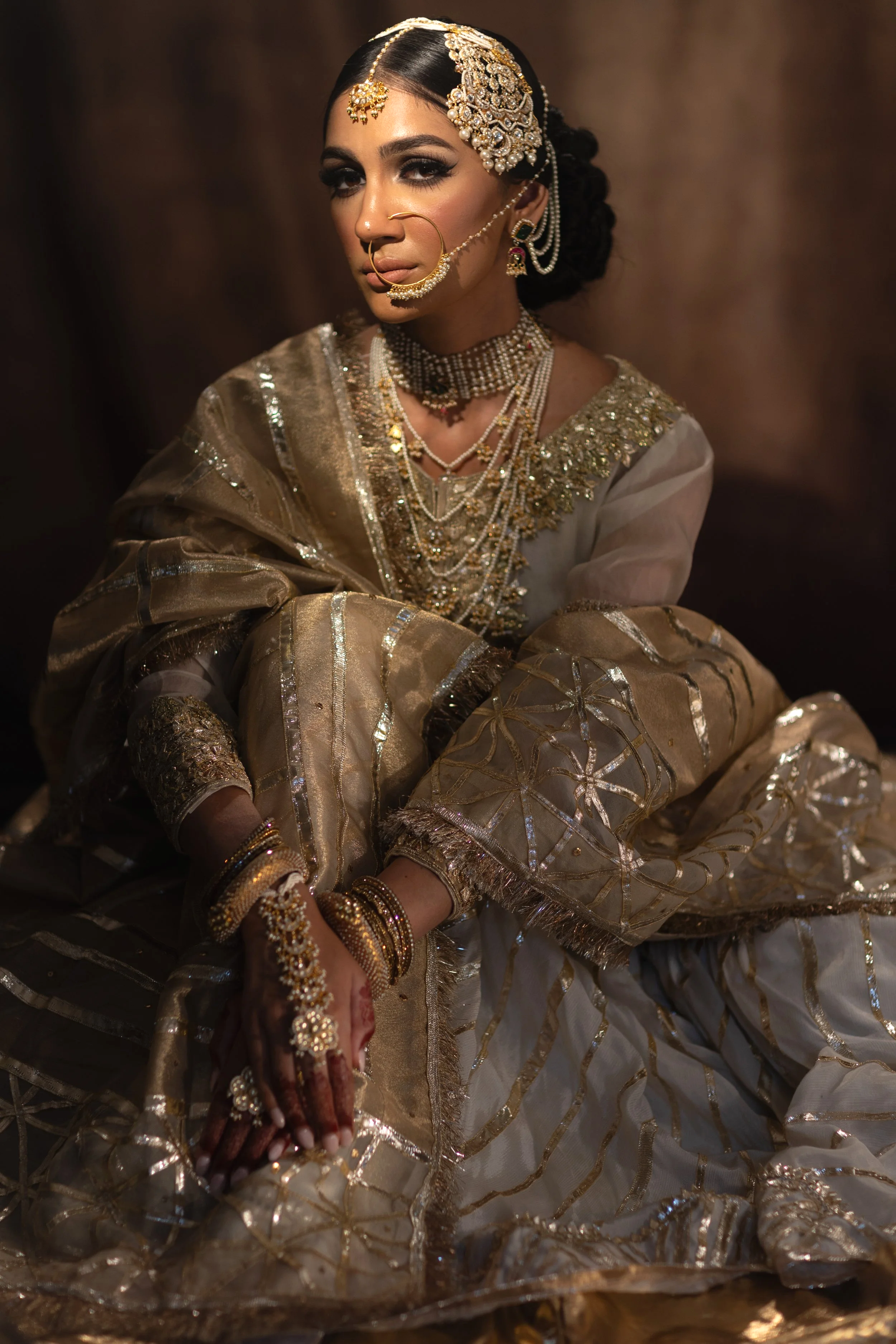 A woman dressed in traditional Indian bridal attire with gold and pearl jewelry, including a head ornament, nose ring, necklaces, bangles, and earrings, sitting with her hands folded.