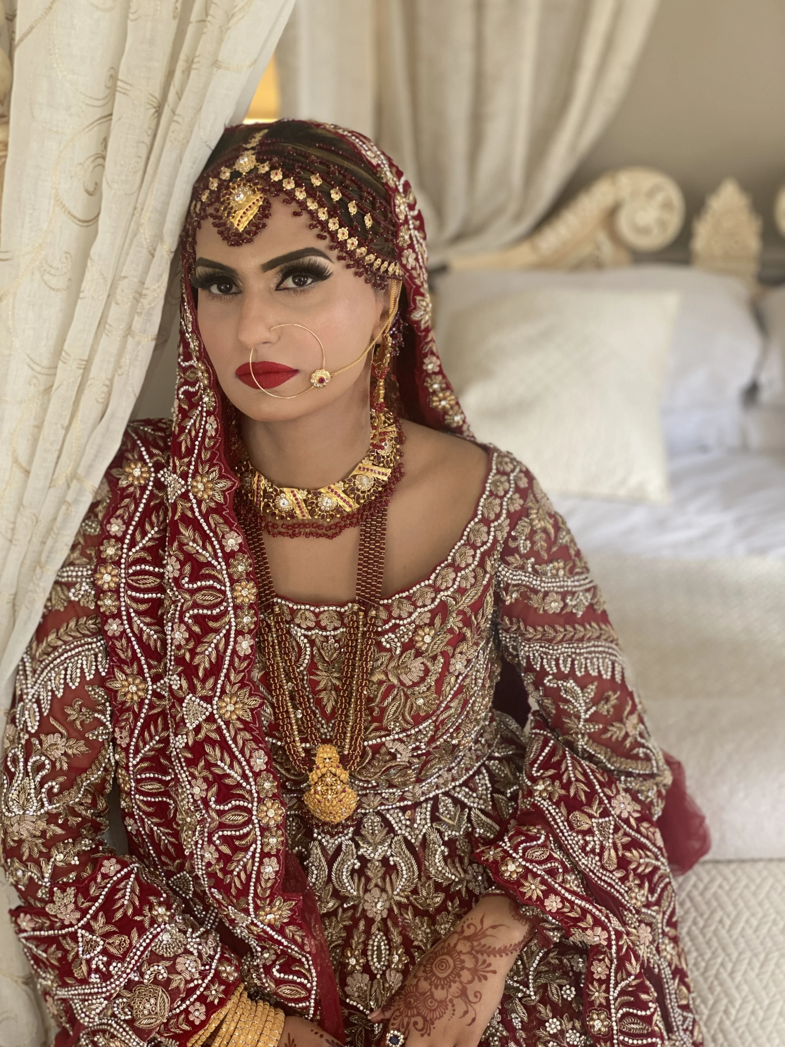 Woman dressed in traditional red and gold bridal attire with jewelry, henna on her hands, and a nose ring, sitting beside a cream-colored curtain.