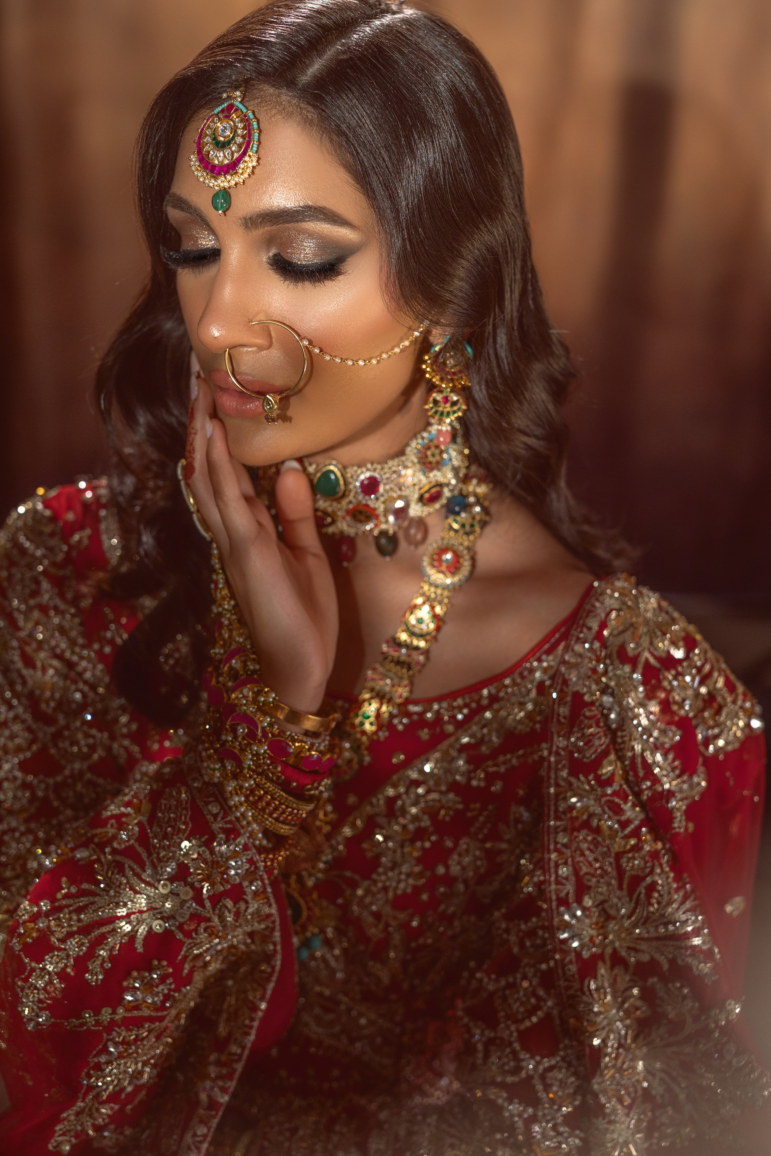 A woman dressed in traditional Indian bridal attire, wearing intricate gold and gemstone jewelry, with her eyes closed and hand gently resting near her face.