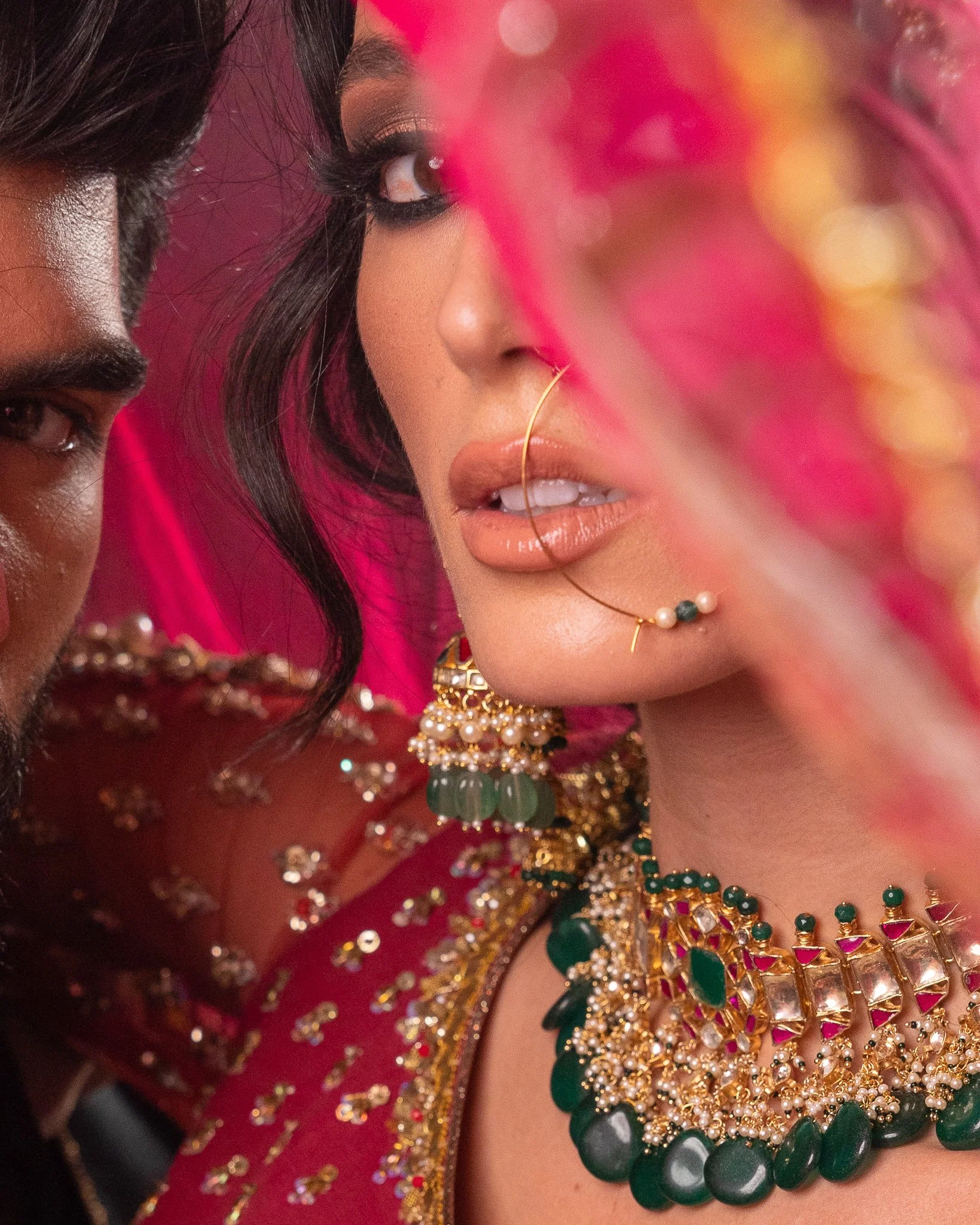 A close-up of a woman in traditional Indian jewelry and clothing, with part of another person's face visible on the left, pink and gold background, and pink fabric partially covering her face.