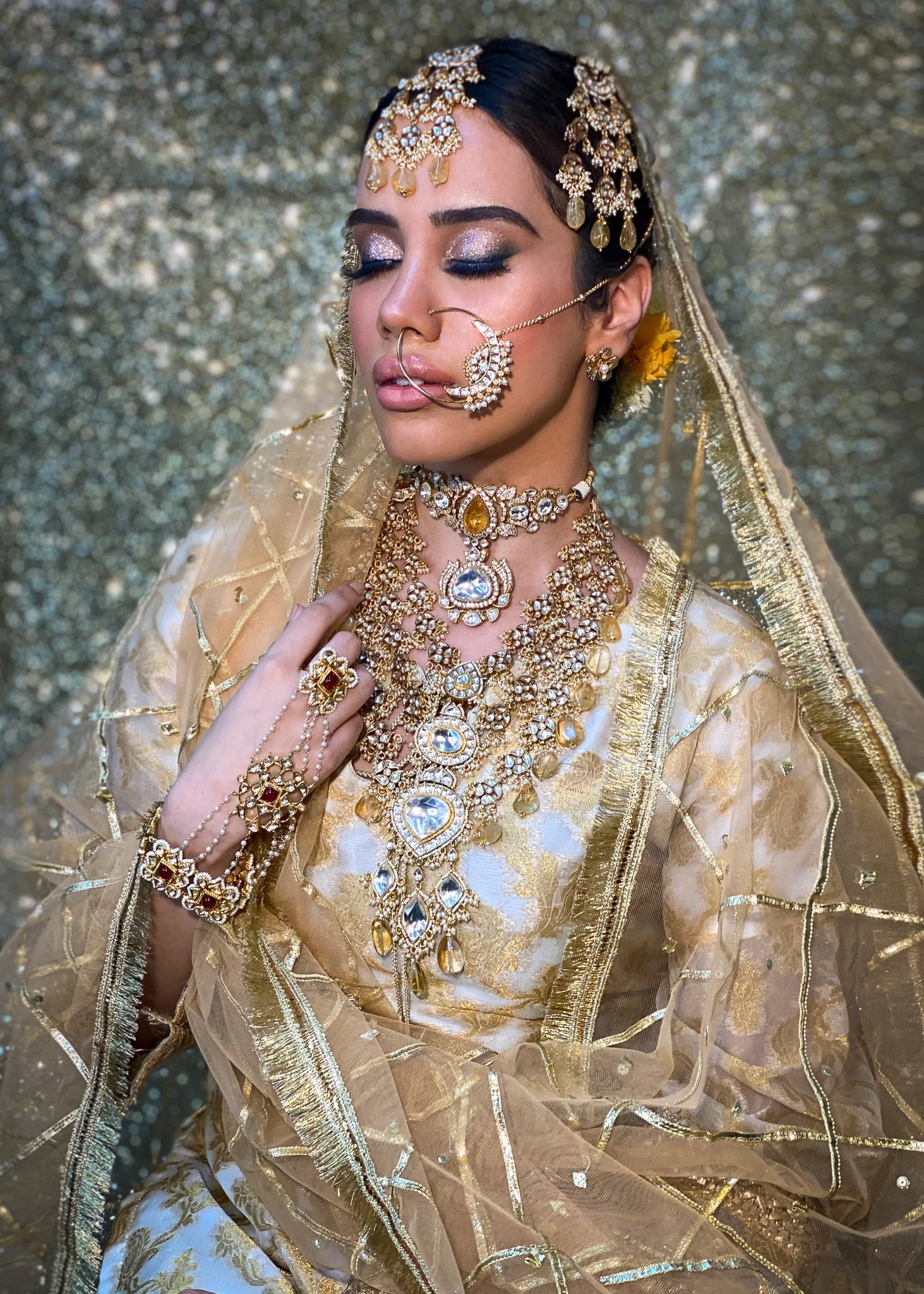 Woman dressed in traditional Indian bridal attire adorned with gold and gemstone jewelry, including necklaces, earrings, a nose ring, and a headpiece, with a golden embroidered veil and shimmering background.