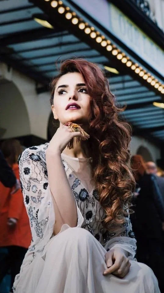 A woman with long, curly red hair sitting in a crowded, indoor public space with a decorative ceiling. She is wearing a white dress with floral embroidery and holding a small purse or clutch. She has a thoughtful expression on her face.