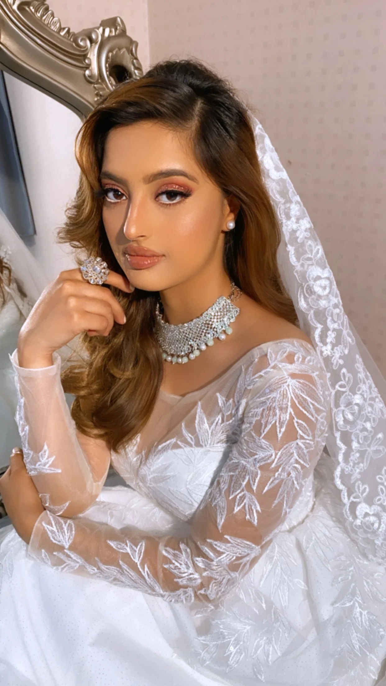 A bride in a white lace wedding dress with floral patterns, wearing pearl earrings, a pearl and rhinestone necklace, and a large ring. She has long, wavy brown hair, makeup with pink eyeshadow, and a subtle lip color. She is posed indoors against a mirror with a decorative frame.