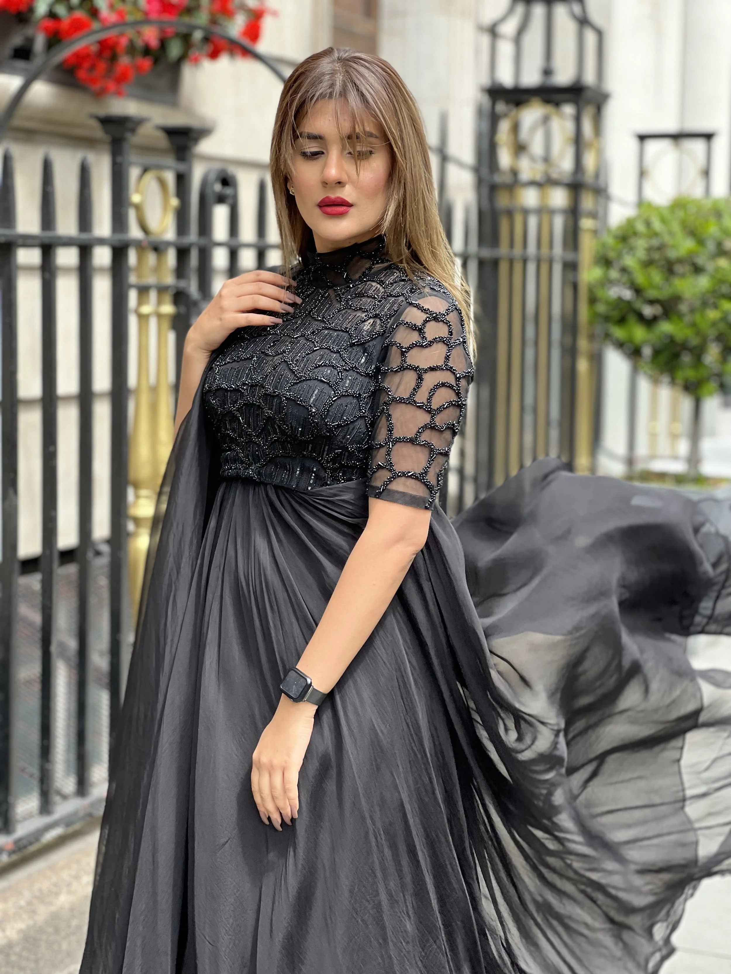 Woman in black gown with sheer beaded sleeves and flowing skirt, standing outdoors near a decorative iron fence and greenery.