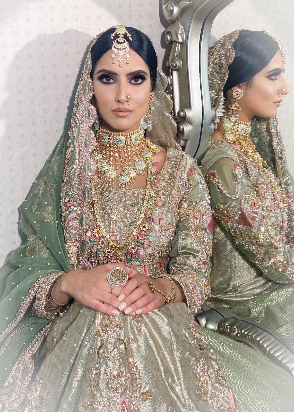 A woman dressed in traditional Indian wedding attire with gold and gemstone jewelry, sitting in front of a mirror.