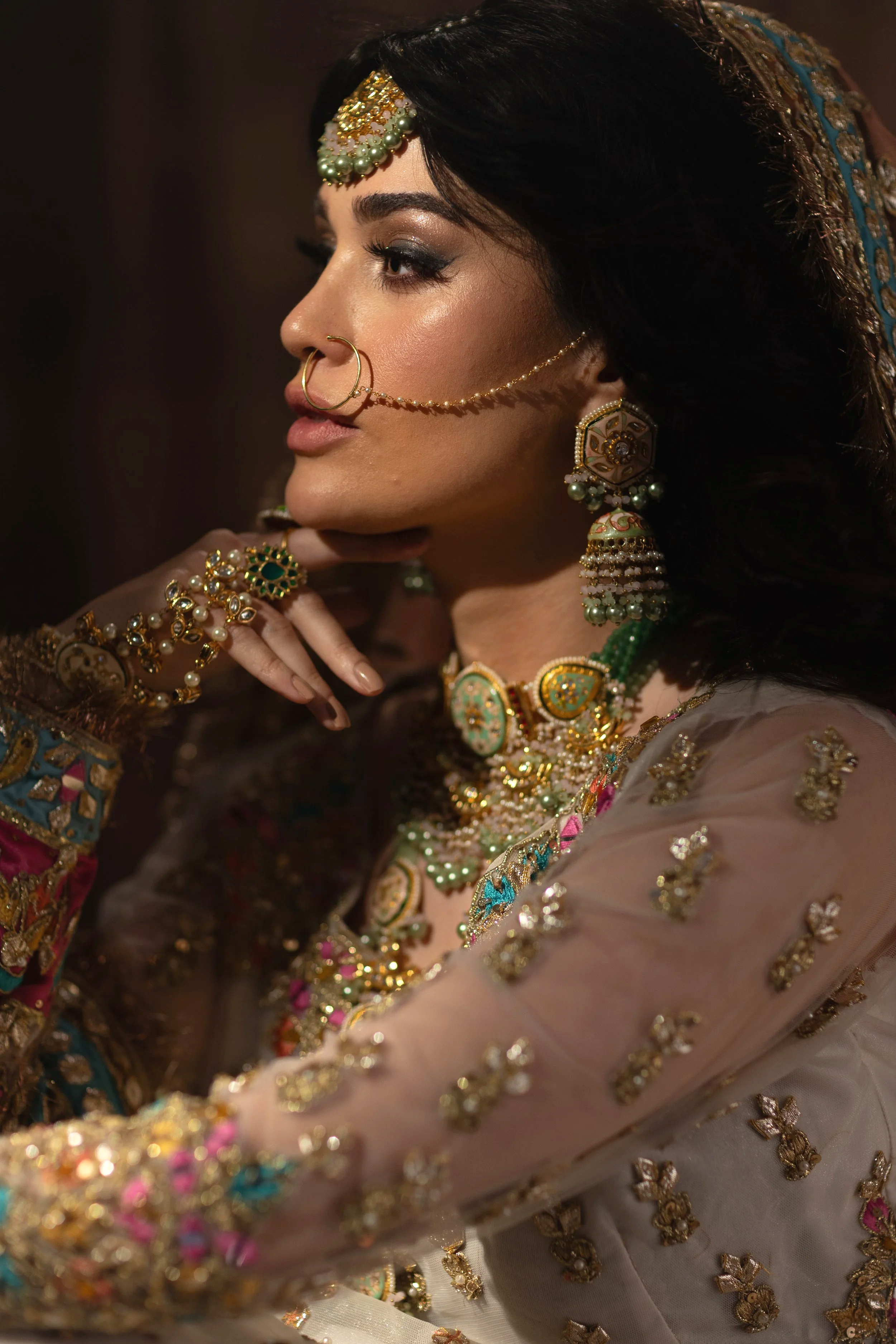 A woman dressed in traditional Indian attire and jewelry, wearing ornate earrings, necklaces, bangles, and a nose ring, with her hand resting on her chin.