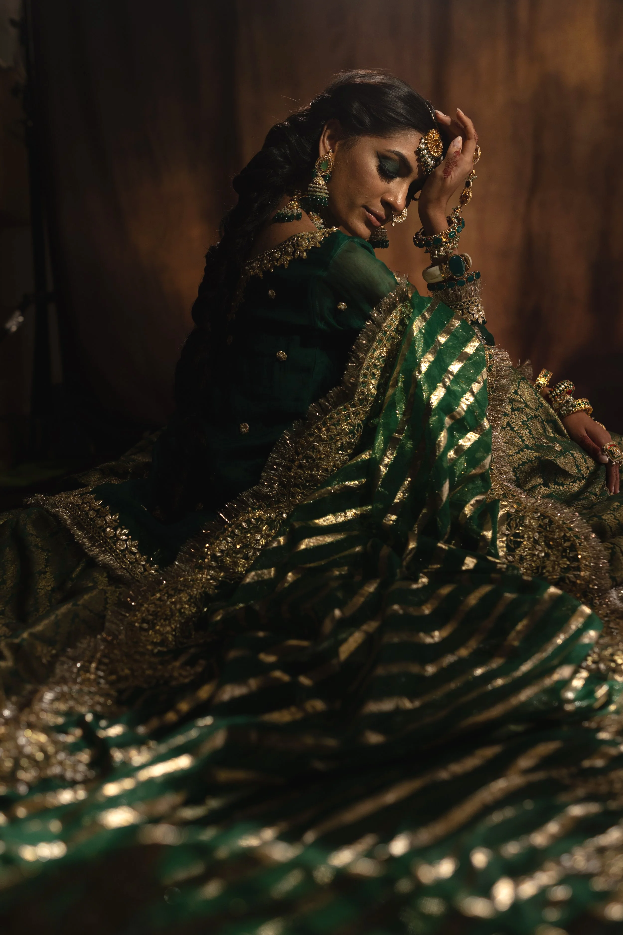 A woman dressed in traditional Indian green and gold attire, adorned with jewelry, sitting against a dark background with her eyes closed and hand resting on her forehead.