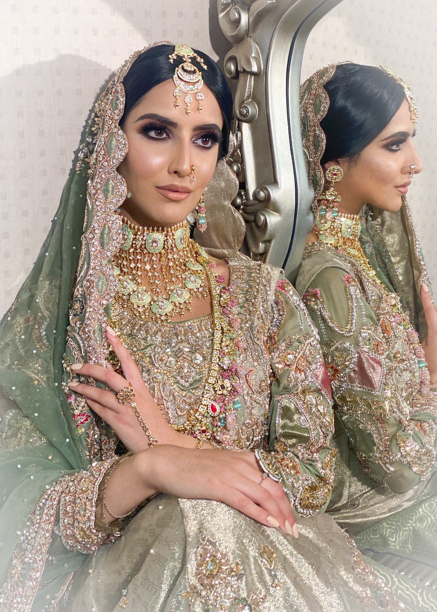 A woman dressed in traditional bridal attire, wearing intricate gold jewelry, including a headpiece, earrings, necklace, and bangles, seated in front of a mirror with her reflection visible.