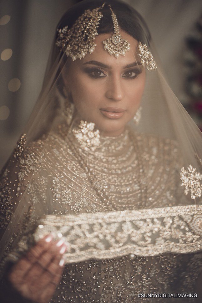 Close-up of a bride dressed in traditional Indian bridal attire with intricate silver embroidery and jewelry, partially covered by a sheer veil.