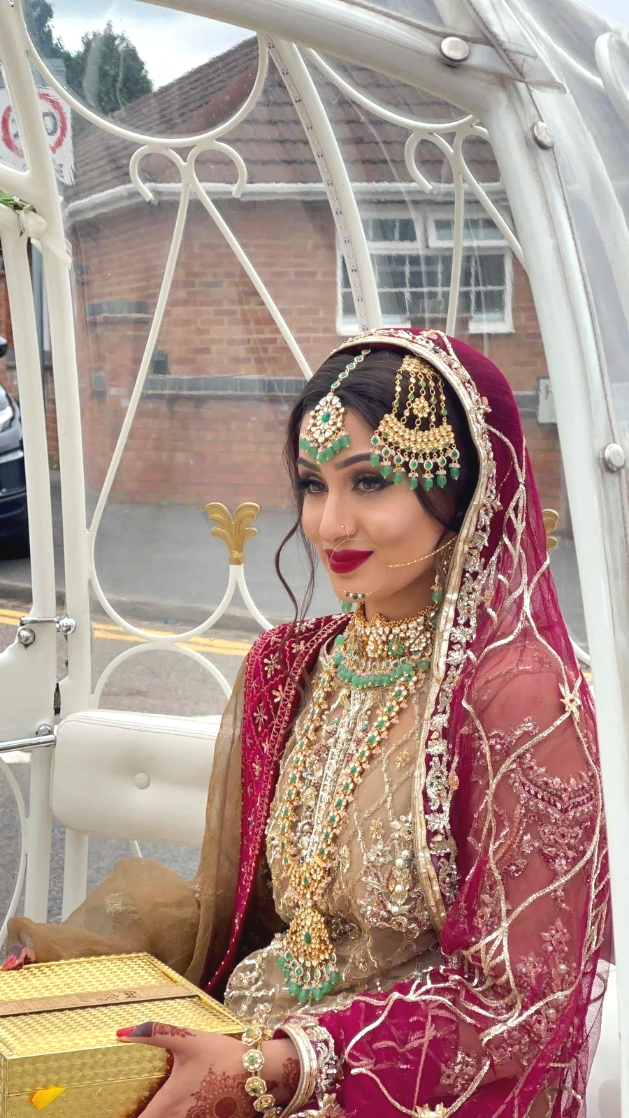 A bride in traditional Indian wedding attire, adorned with an elaborate gold and green jewelry set, sitting inside a white decorative cart, with a brick house in the background.